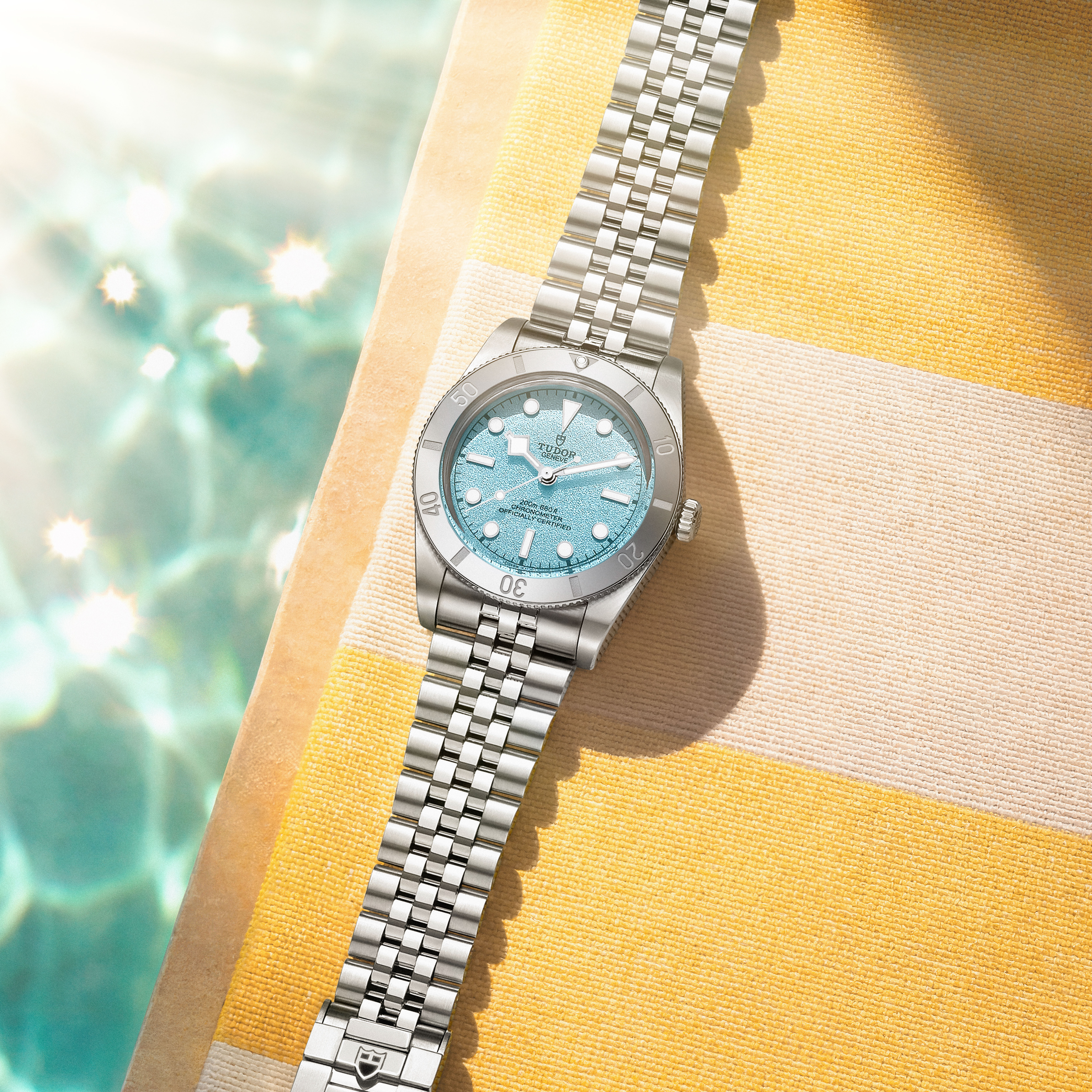 A silver watch with a light blue face and bezel rests on a yellow and white striped towel next to a swimming pool.