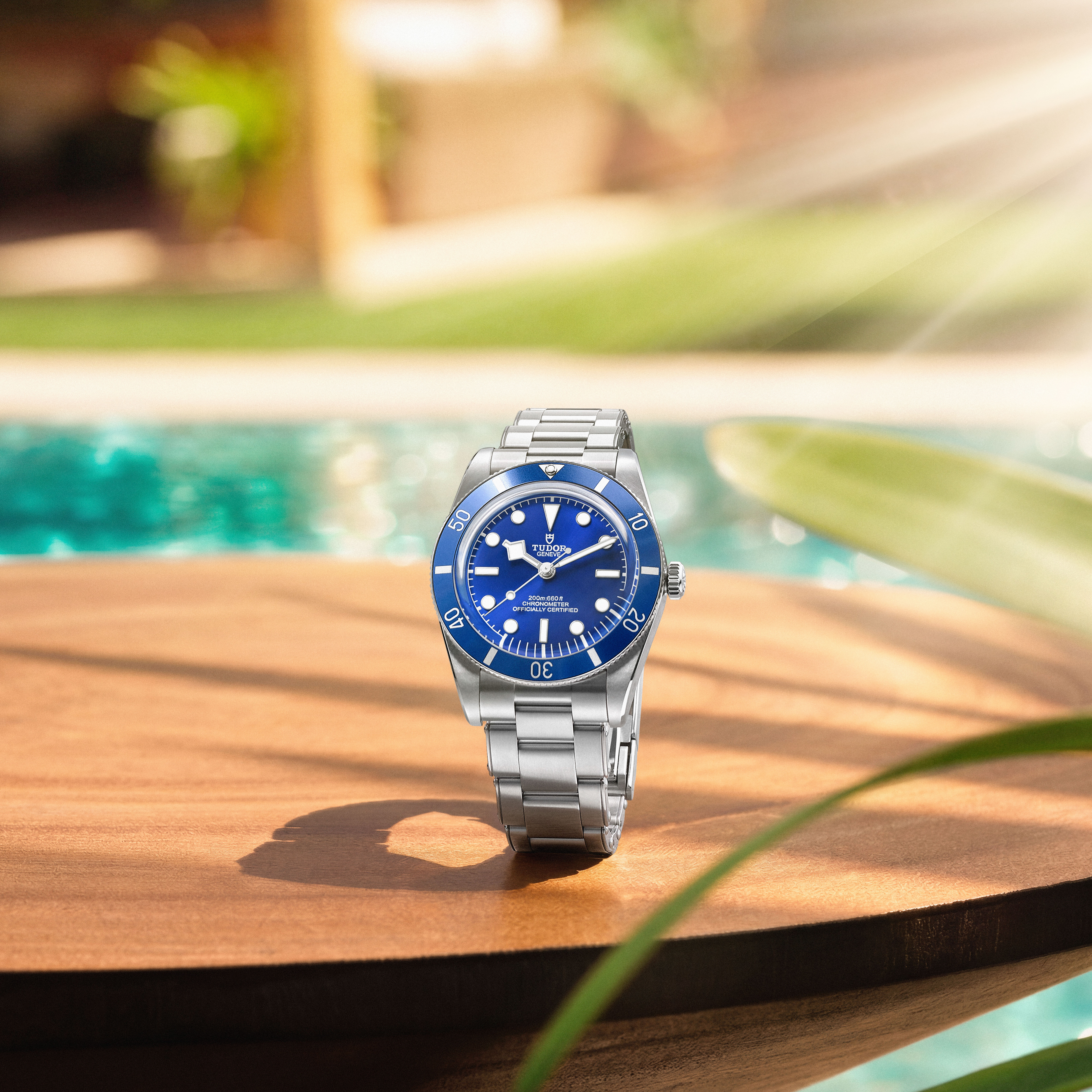 A silver watch with a blue face and bezel sits on a wooden table by a swimming pool.