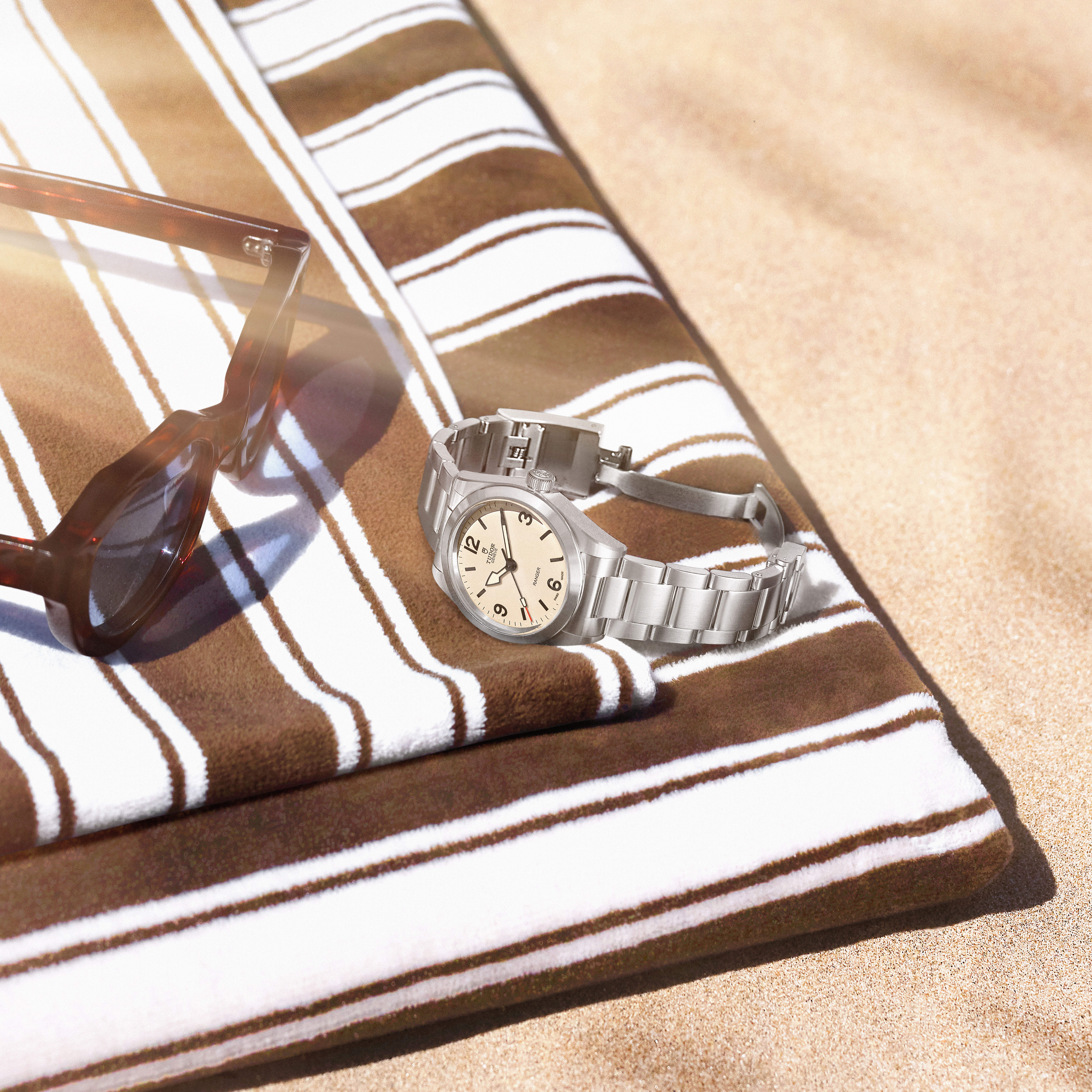 A silver watch with a cream dial and brown sunglasses rest on a striped brown and white towel on a sandy beach.