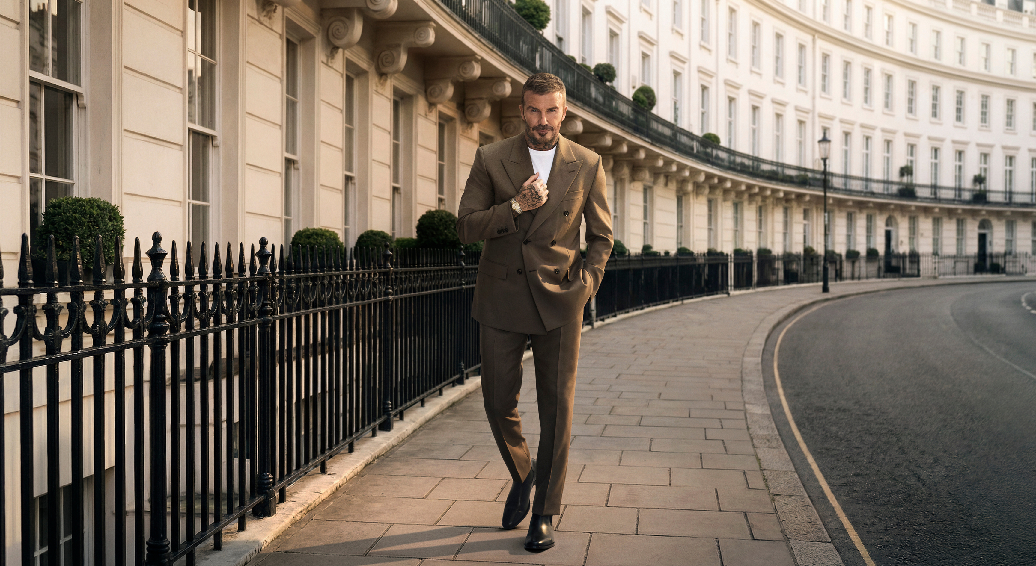 David Beckham in a brown suit walking on a sidewalk next to a black fence and a curved white building.