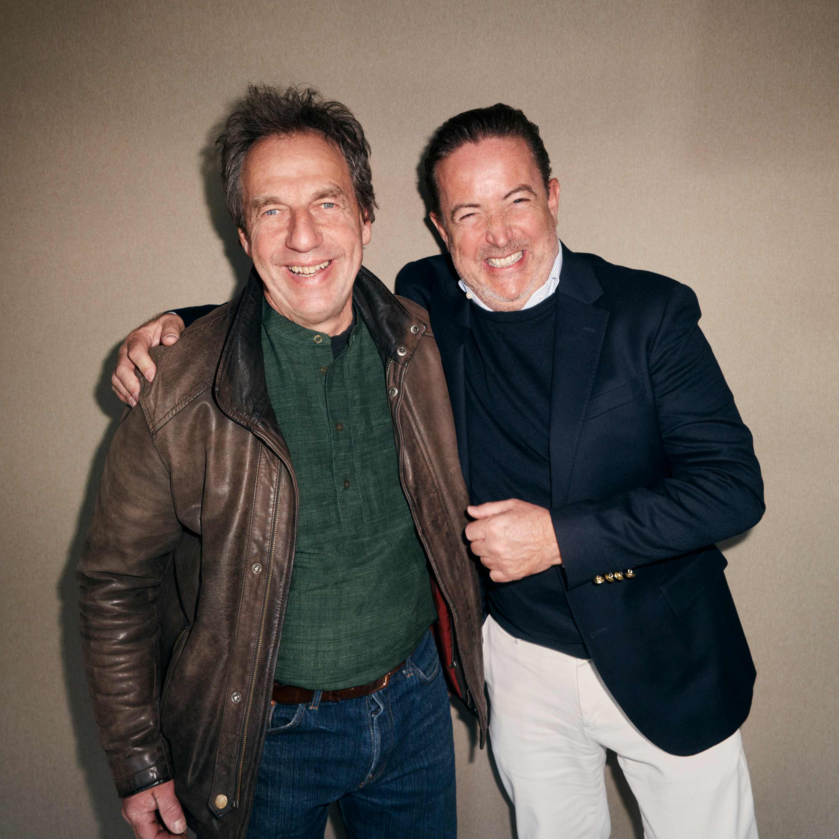 Two smiling men, one in a brown leather jacket and the other in a navy blazer, stand with arms around each other.