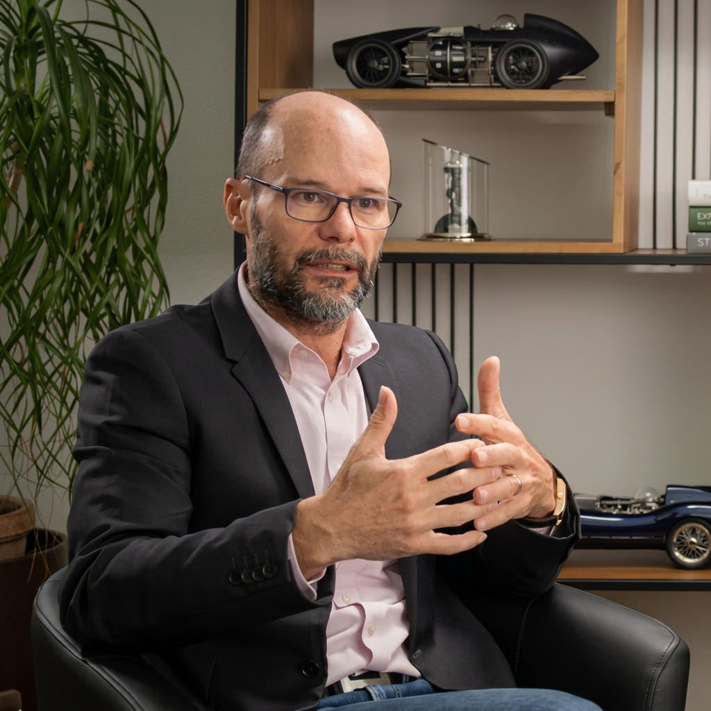 A man with glasses and a beard gestures while sitting in a black chair, with a shelf of model cars behind him.