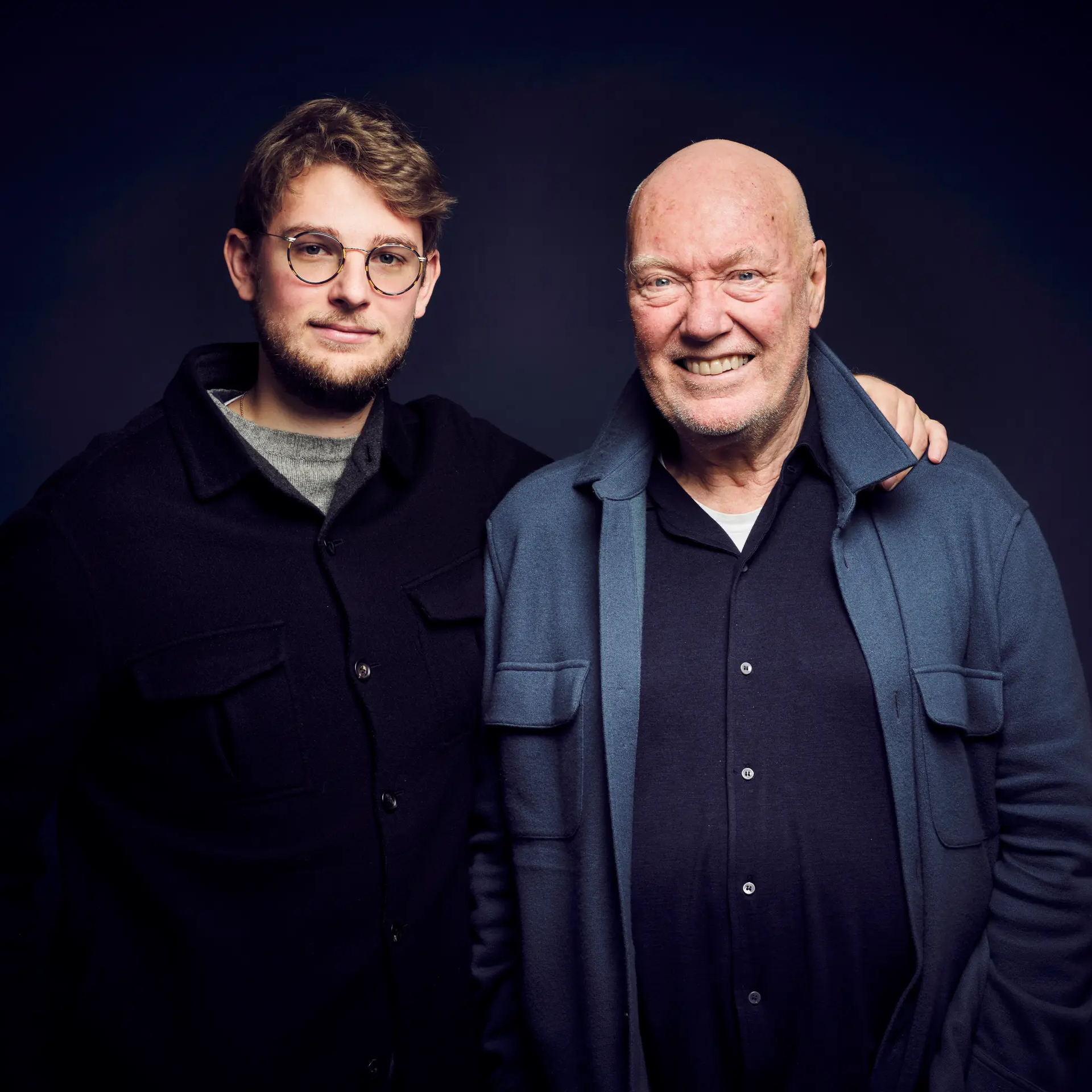 A young man in glasses and a dark jacket stands next to an older bald man in a blue jacket, who has his arm around the younger man.