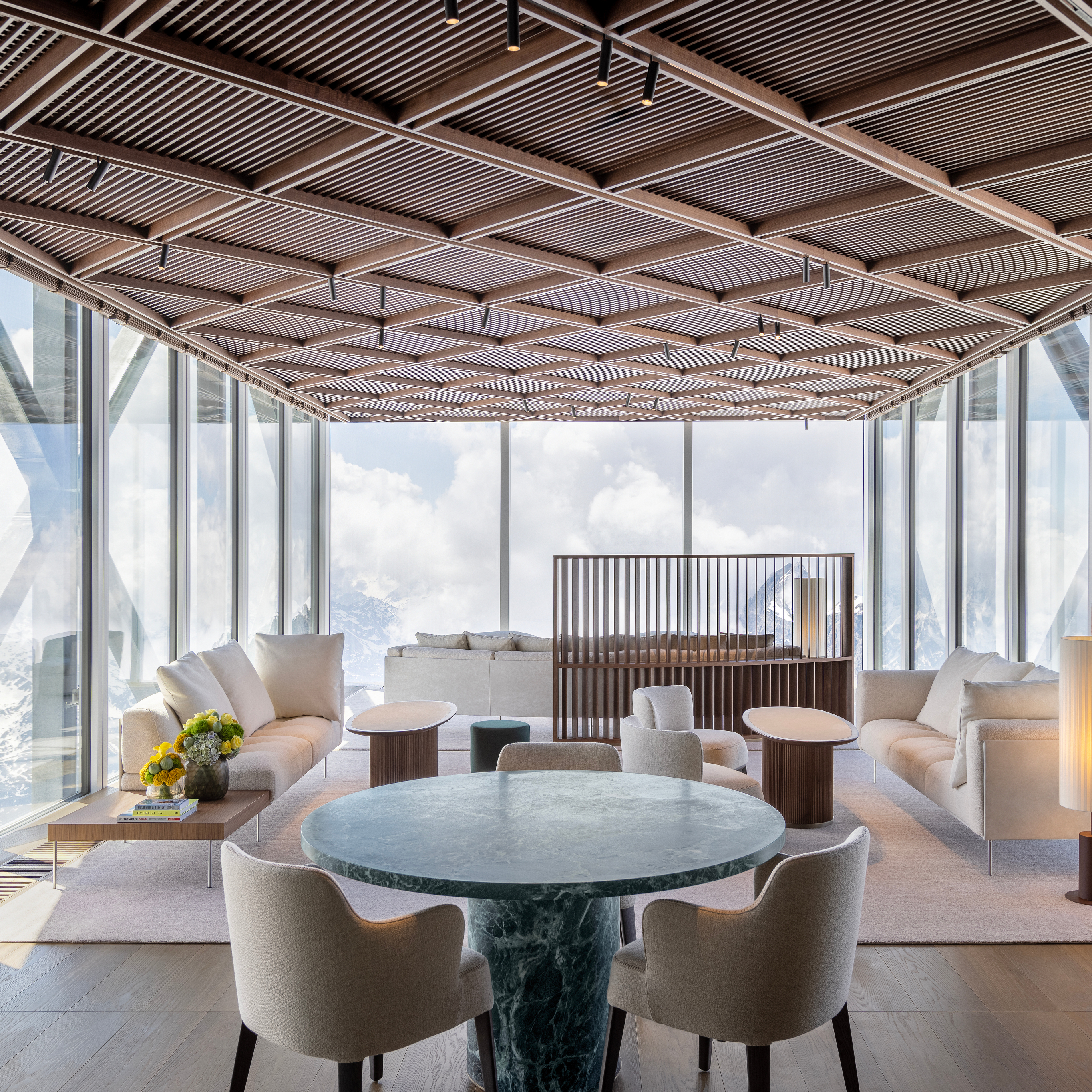 Eine moderne Lounge mit hellen Sofas und Sesseln, einem grünen Marmortisch und einer Lamellendecke aus Holz mit Blick auf schneebedeckte Berge.