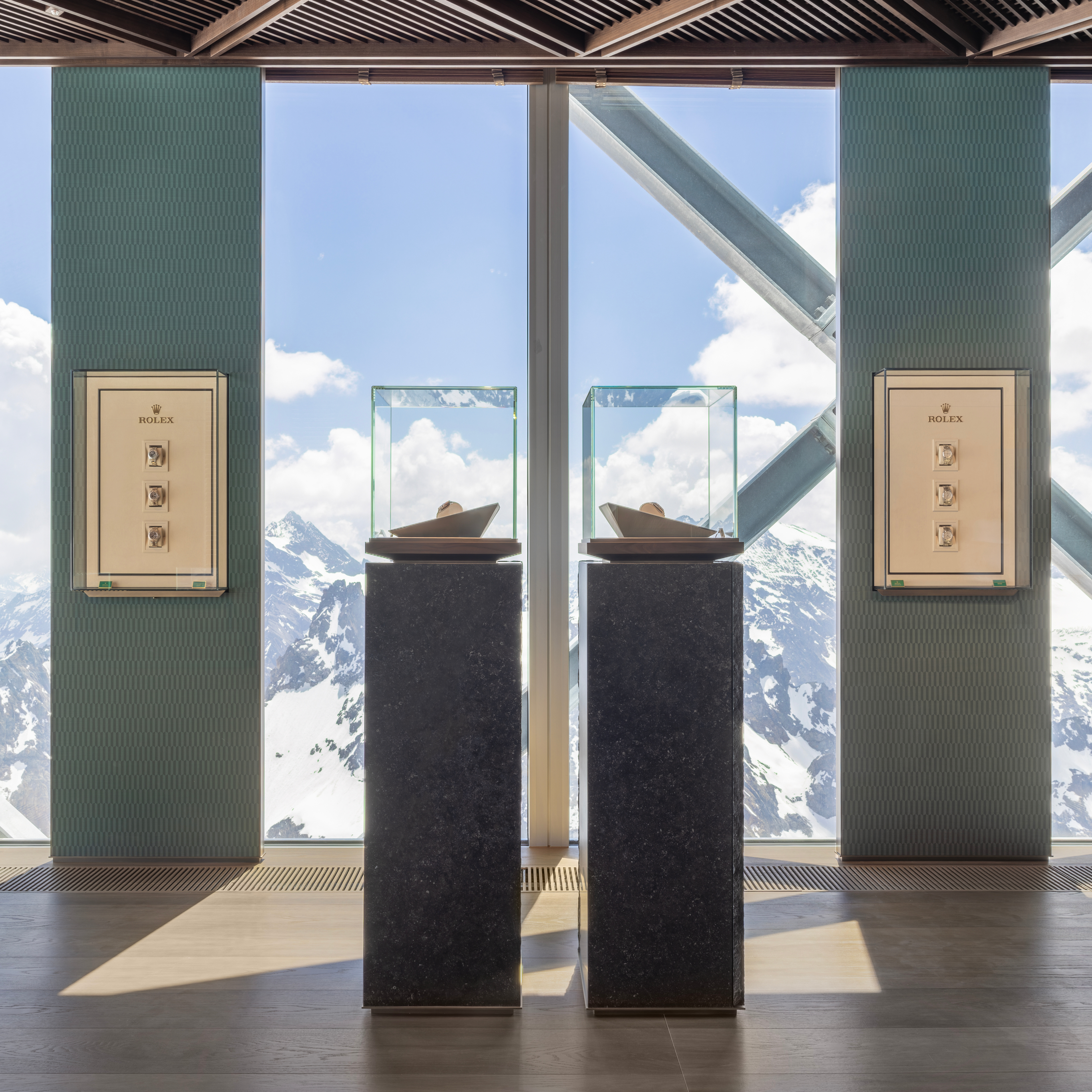 Zwei Rolex-Uhrenvitrinen auf Sockeln vor einem großen Fenster mit Blick auf schneebedeckte Berge.