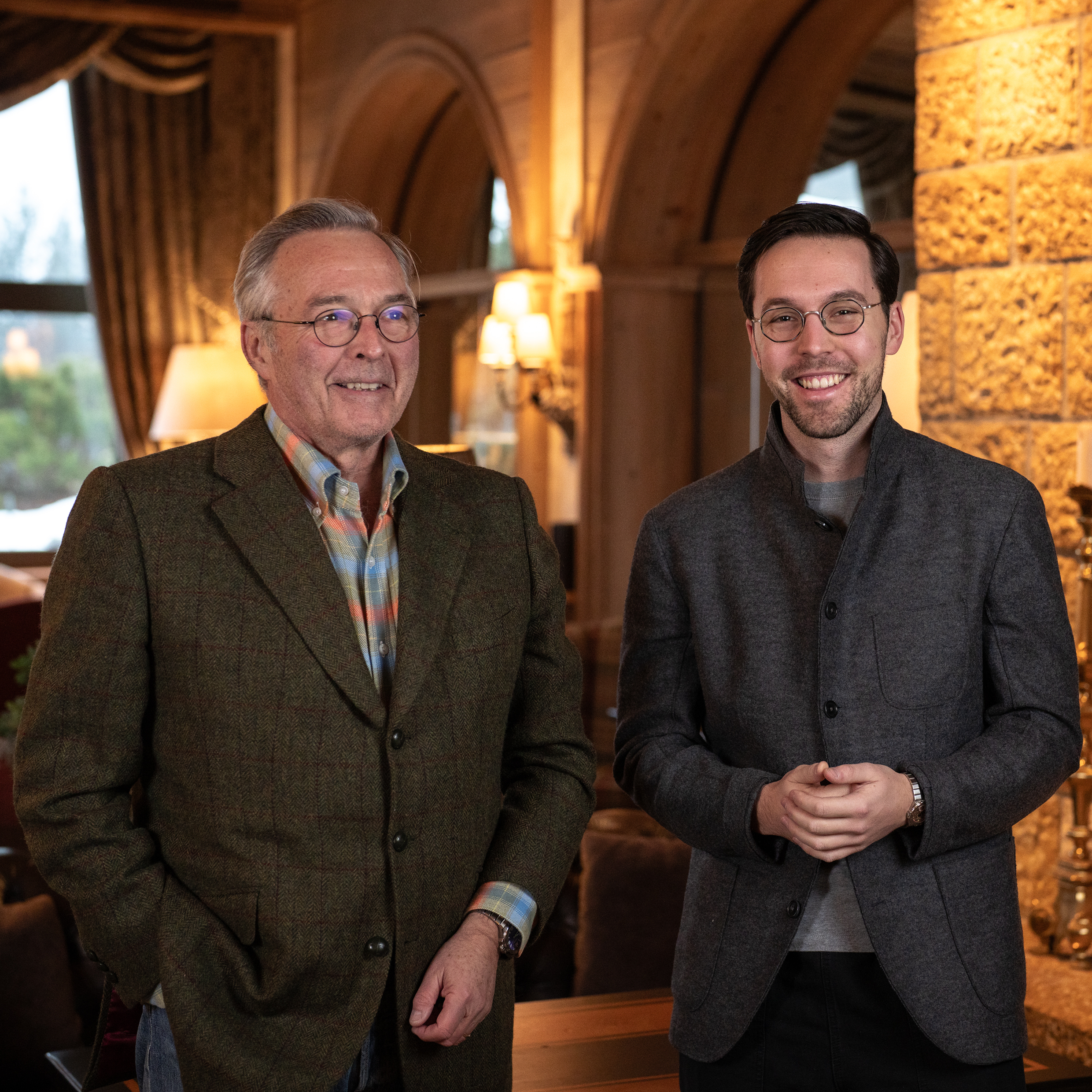 Two men, one older and one younger, stand smiling in a warmly lit, rustic-chic interior.