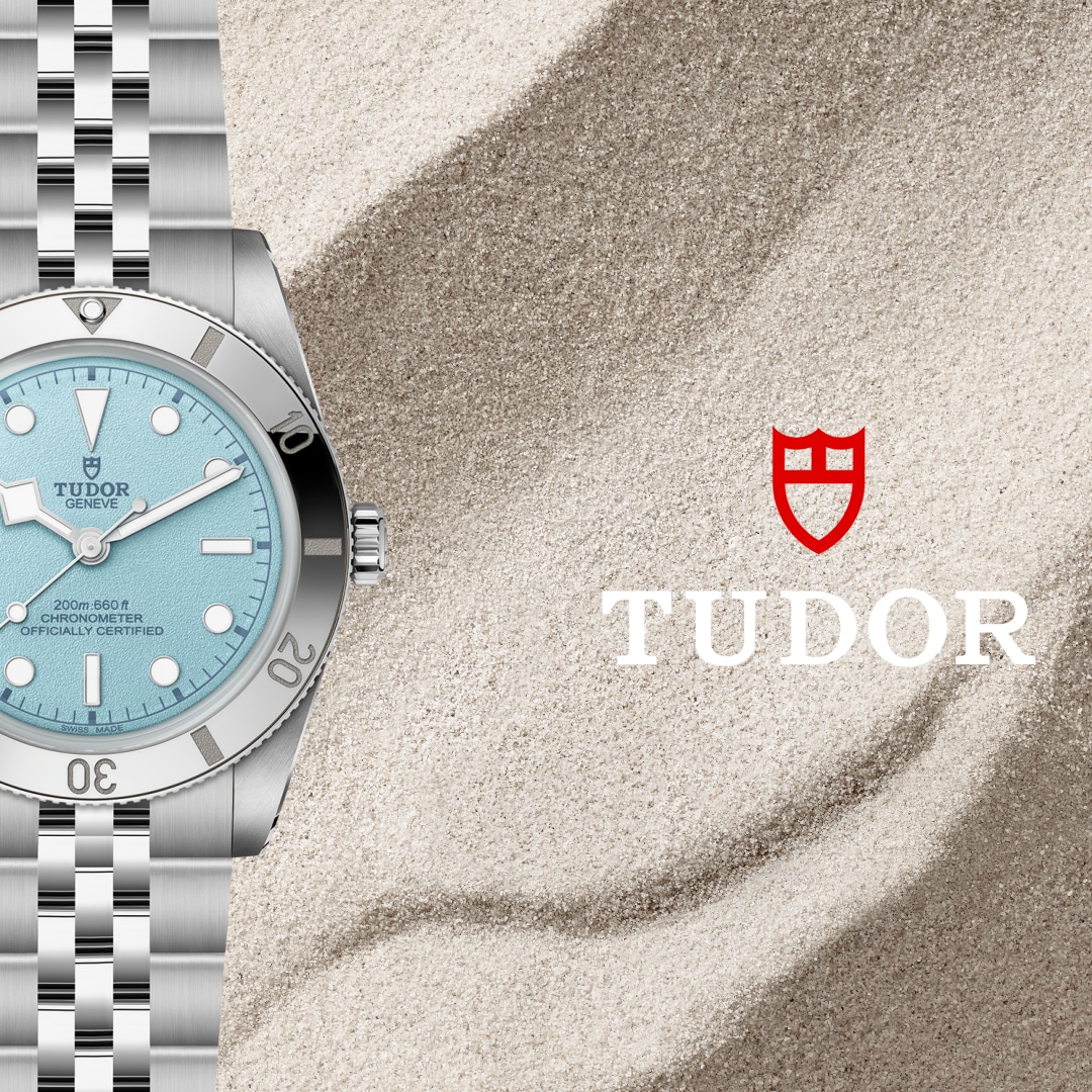 A silver Tudor watch with a light blue face and metal band rests on sand next to the red Tudor logo.