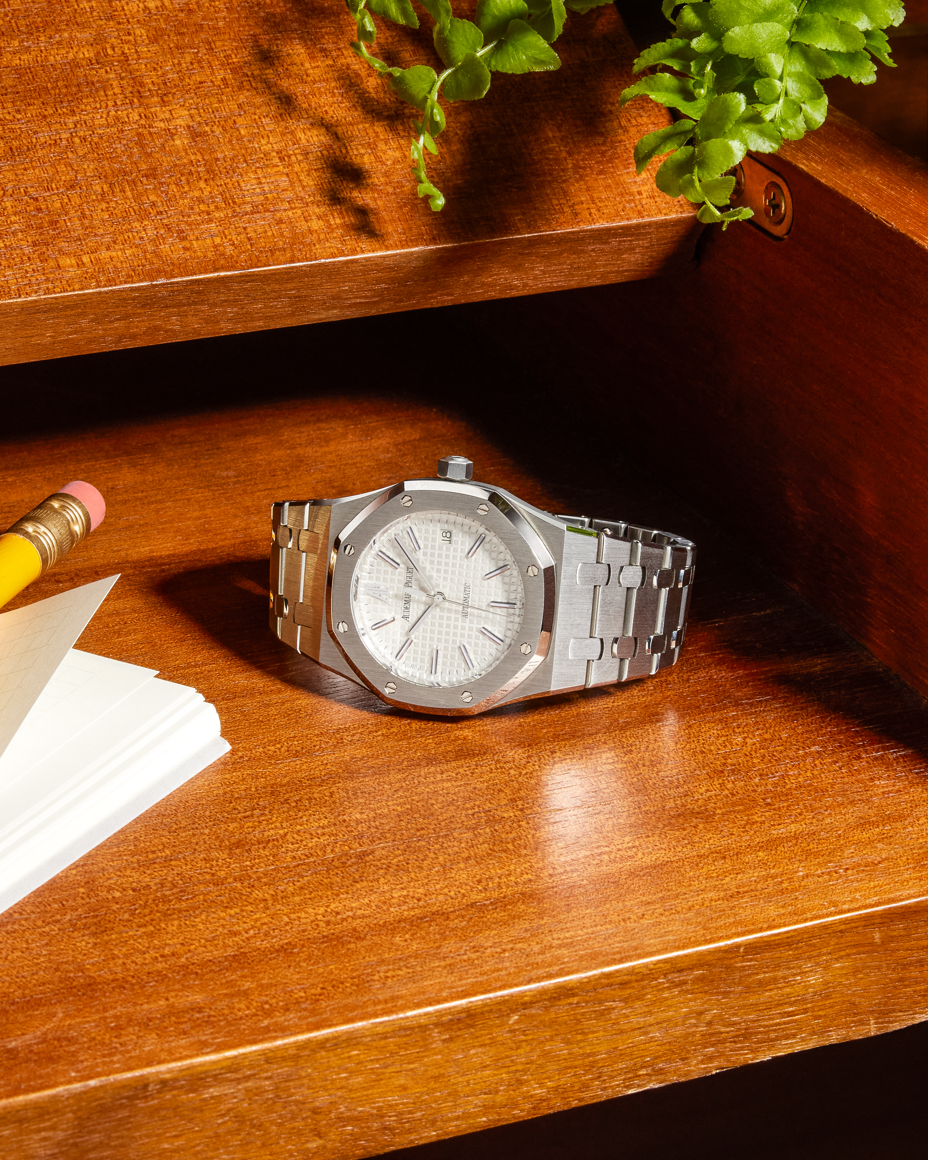 A silver Audemars Piguet Royal Oak watch with a white dial on a wooden desk next to a pencil, notebook, and plant.