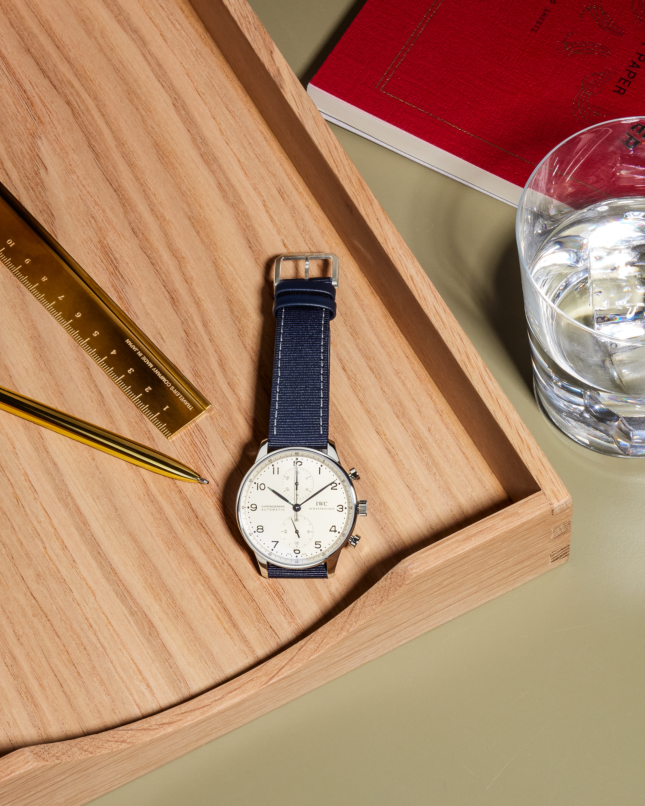 A silver watch with a blue strap, a gold pen, and a ruler on a wooden tray next to a glass of water.