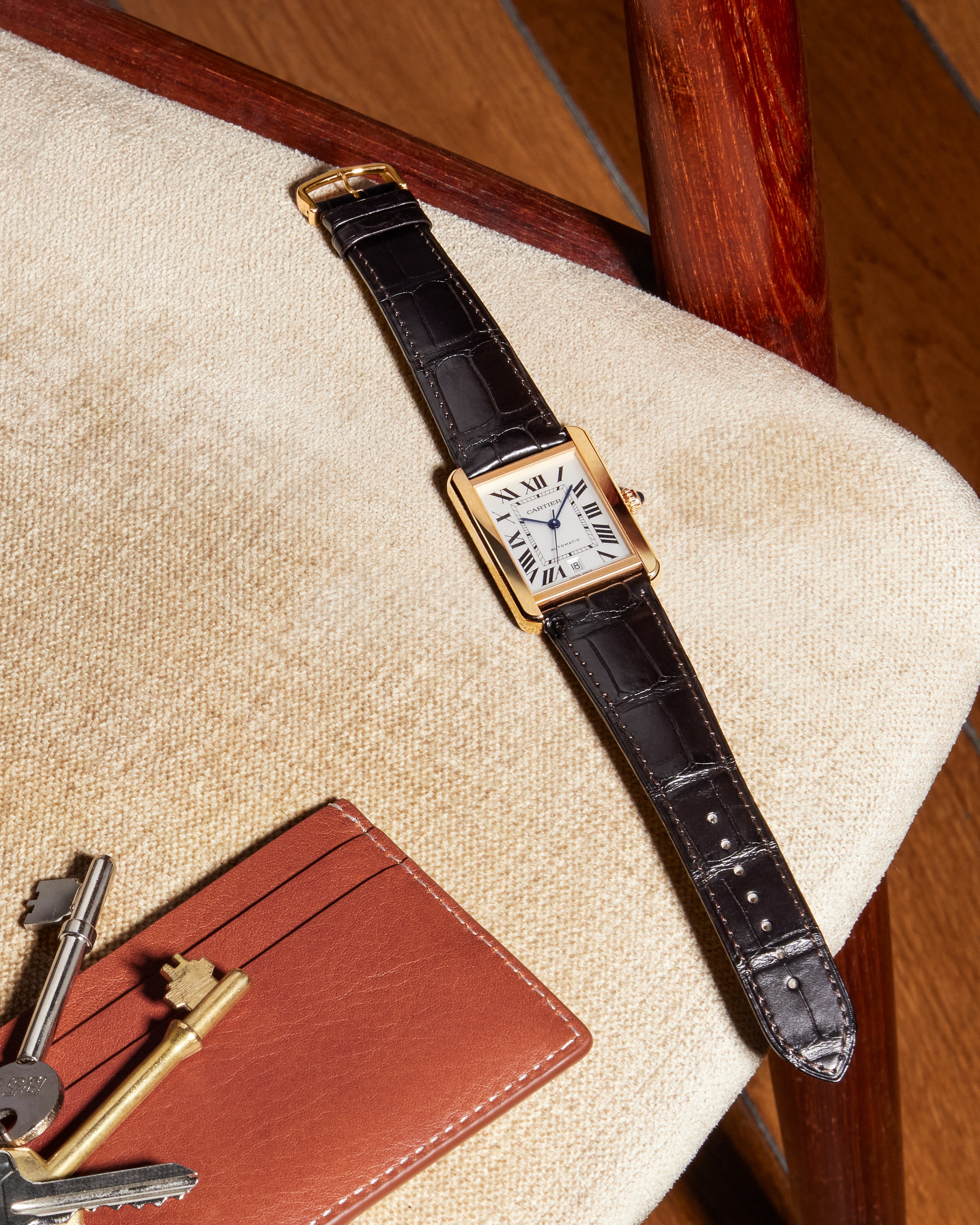 A gold rectangular watch with a black leather strap, keys, and a brown cardholder on a cream chair.