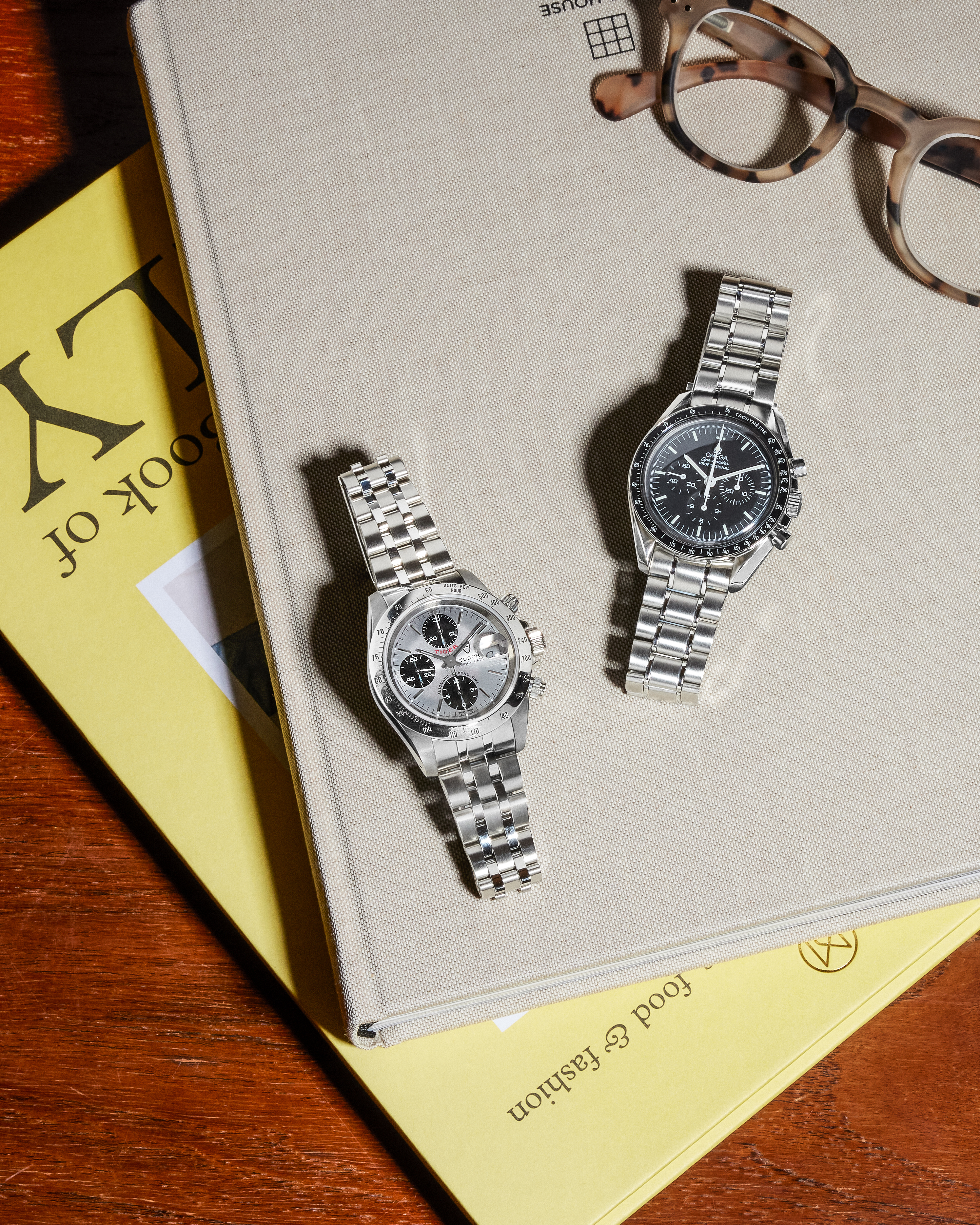 Two silver chronographs with metal bands, one with a black face and one with a silver face, on books.