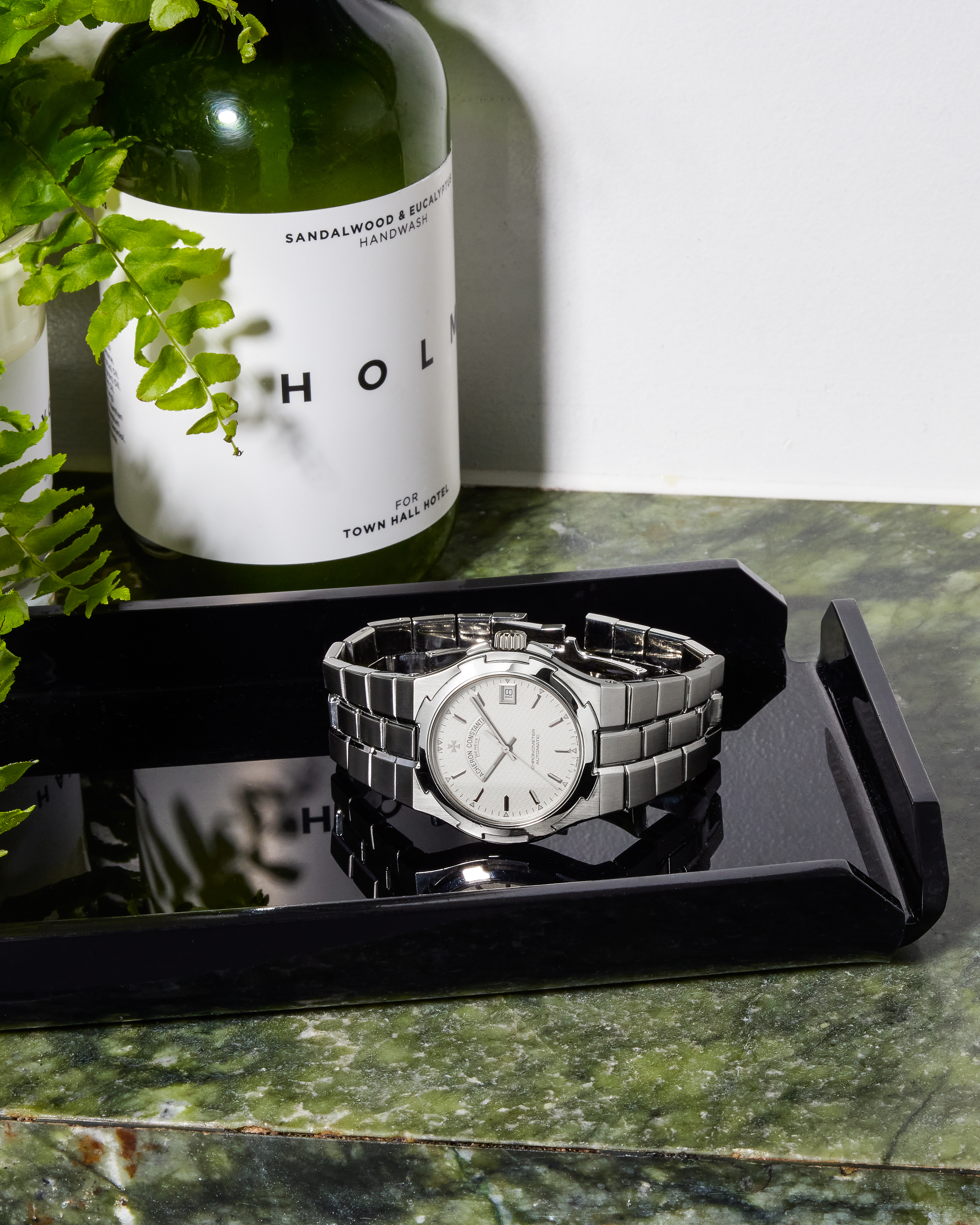 A silver watch with a white face and metal band rests on a black tray next to a bottle of handwash and green plant.