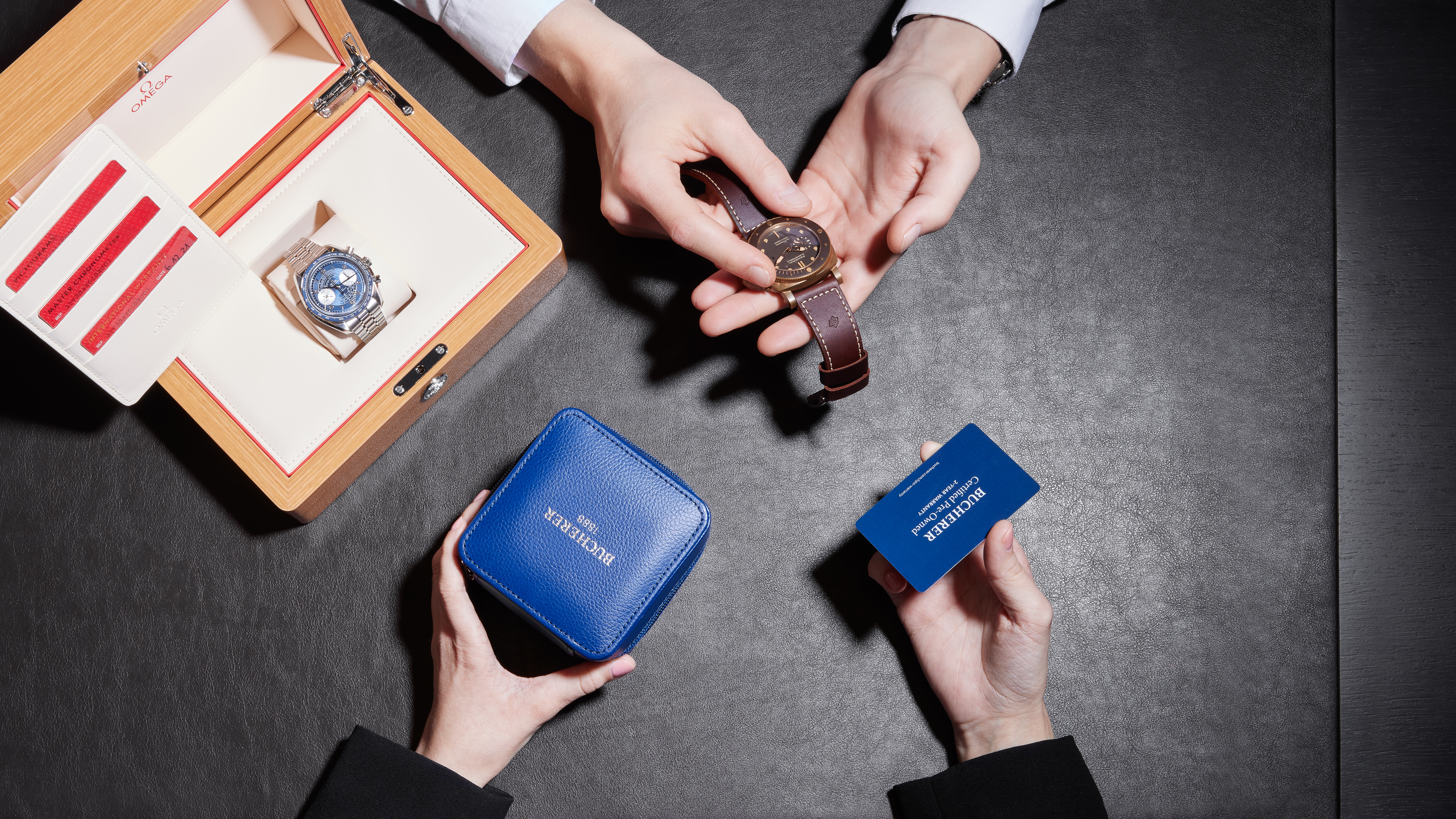 Two people examining luxury watches and a warranty card on a dark surface.