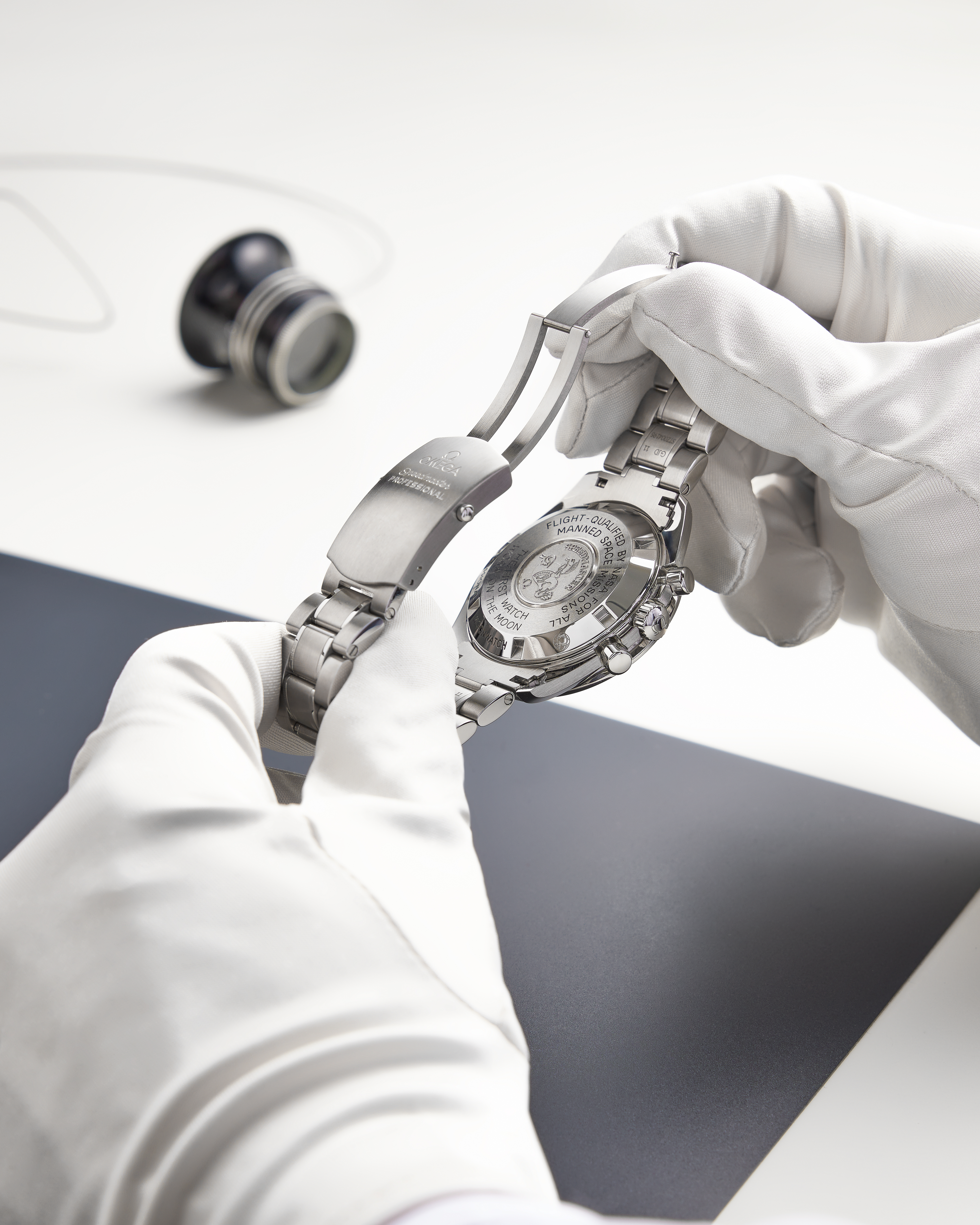 Behandschuhte Hände halten eine silberne Omega Speedmaster-Uhr, deren gravierter Gehäuseboden und Verschluss sichtbar sind.