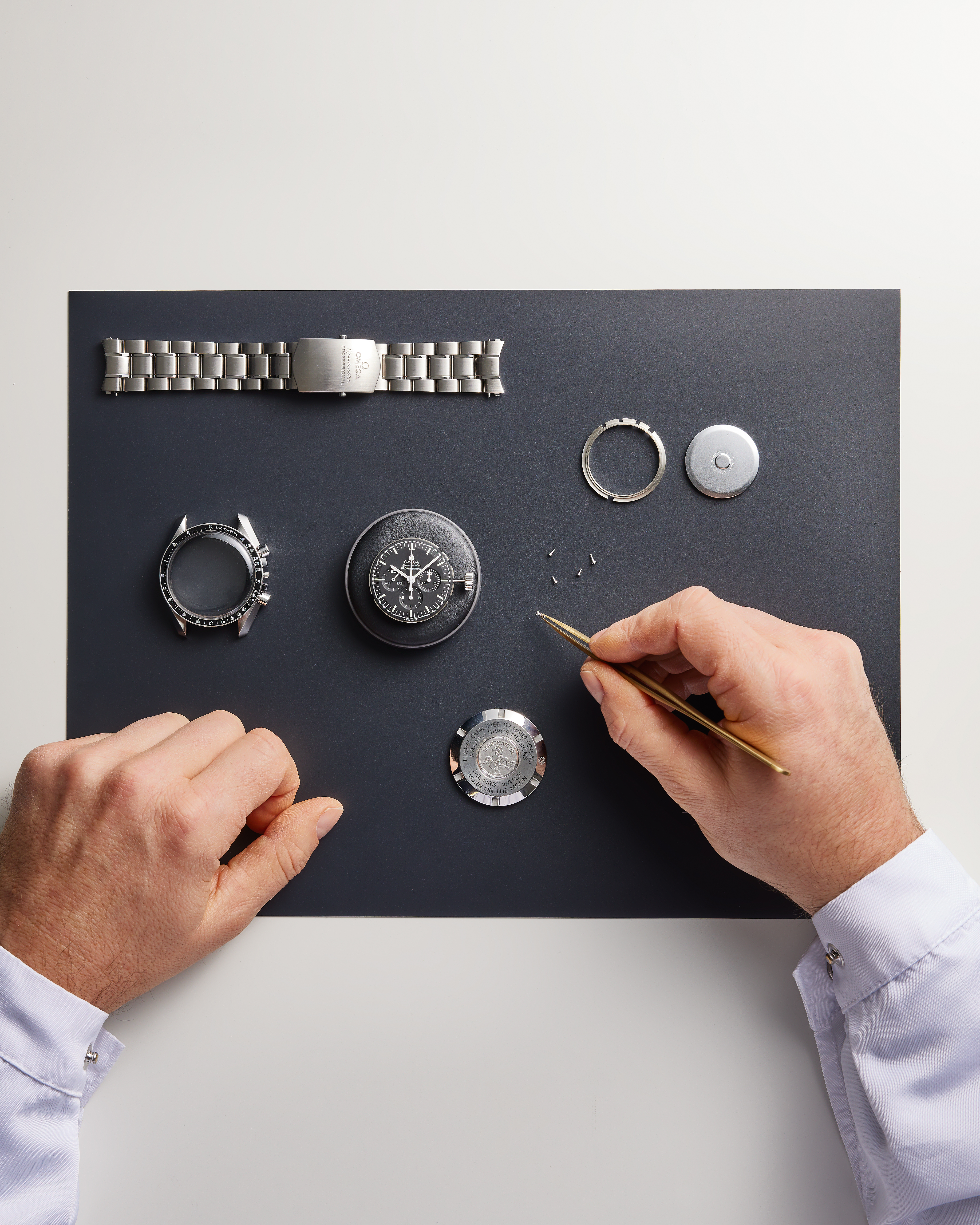 A watchmaker's hands assemble a watch from its components, including the case, movement, and metal bracelet, on a dark mat.