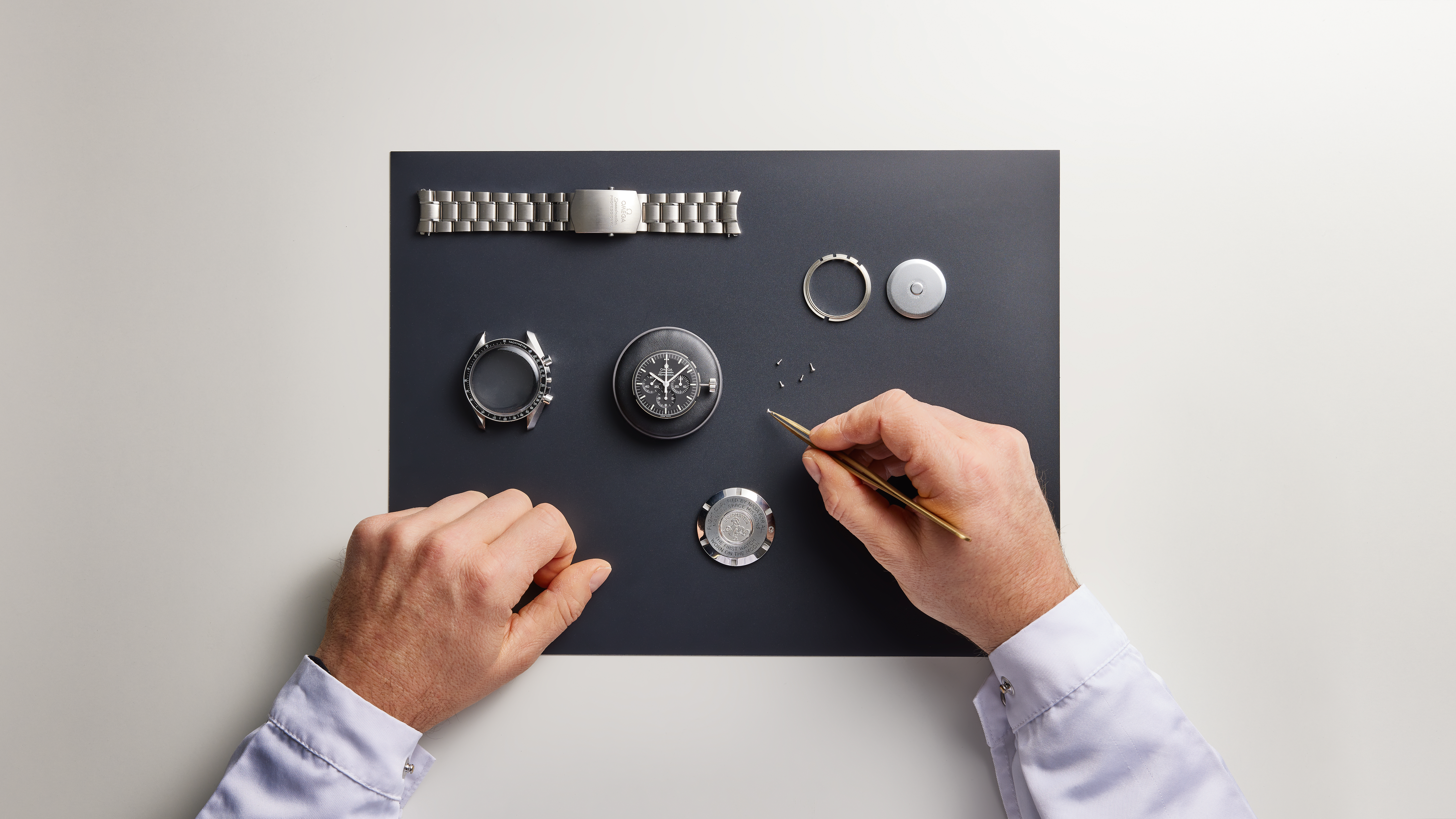 Hands assembling a watch, with components like the watch face, bezel, case, and metal strap laid out on a dark mat.