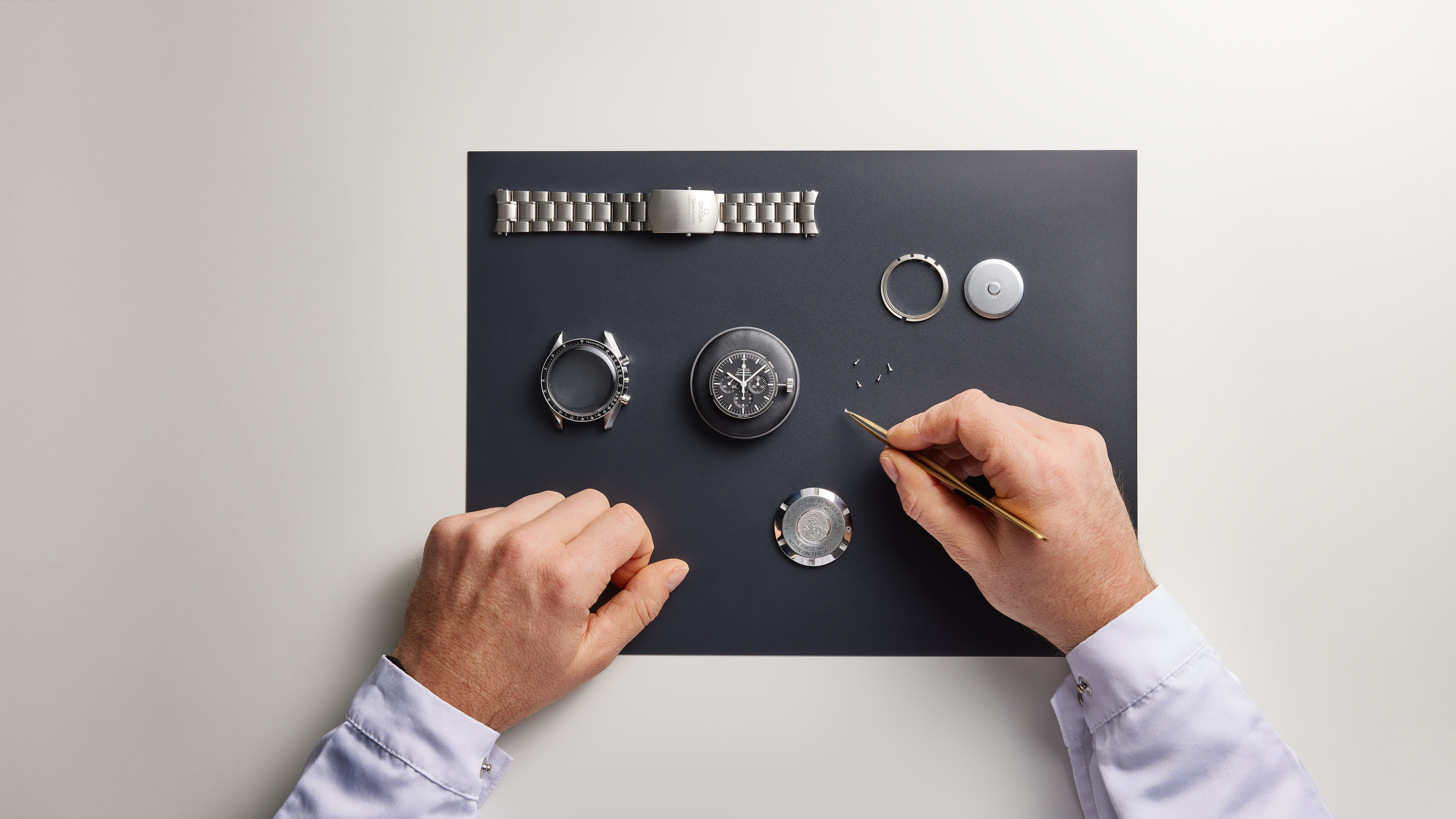 A watchmaker's hands assemble a watch from its components on a black mat.