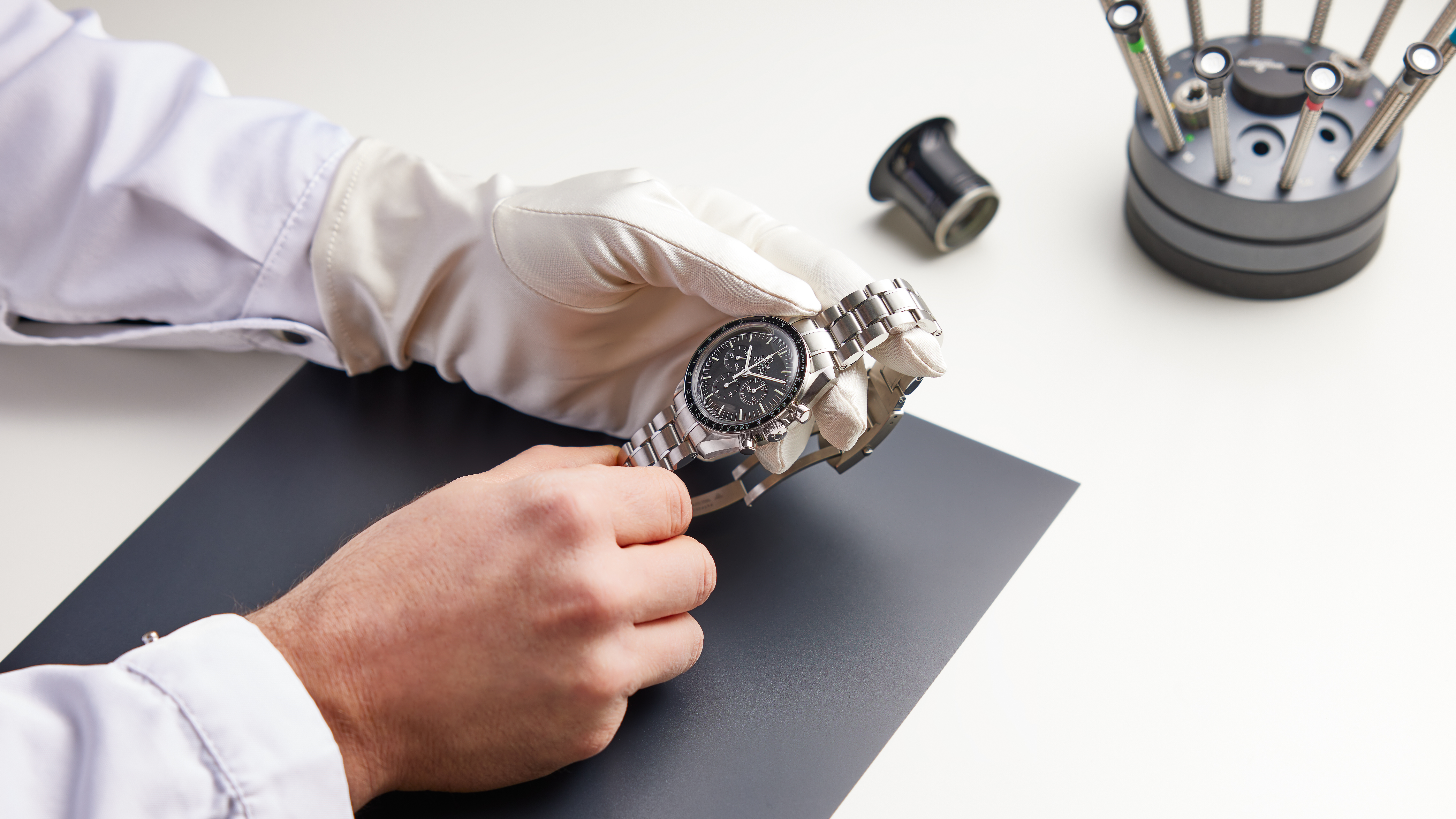 A gloved hand holding a silver watch with a black face, next to a black mat and watchmaking tools.