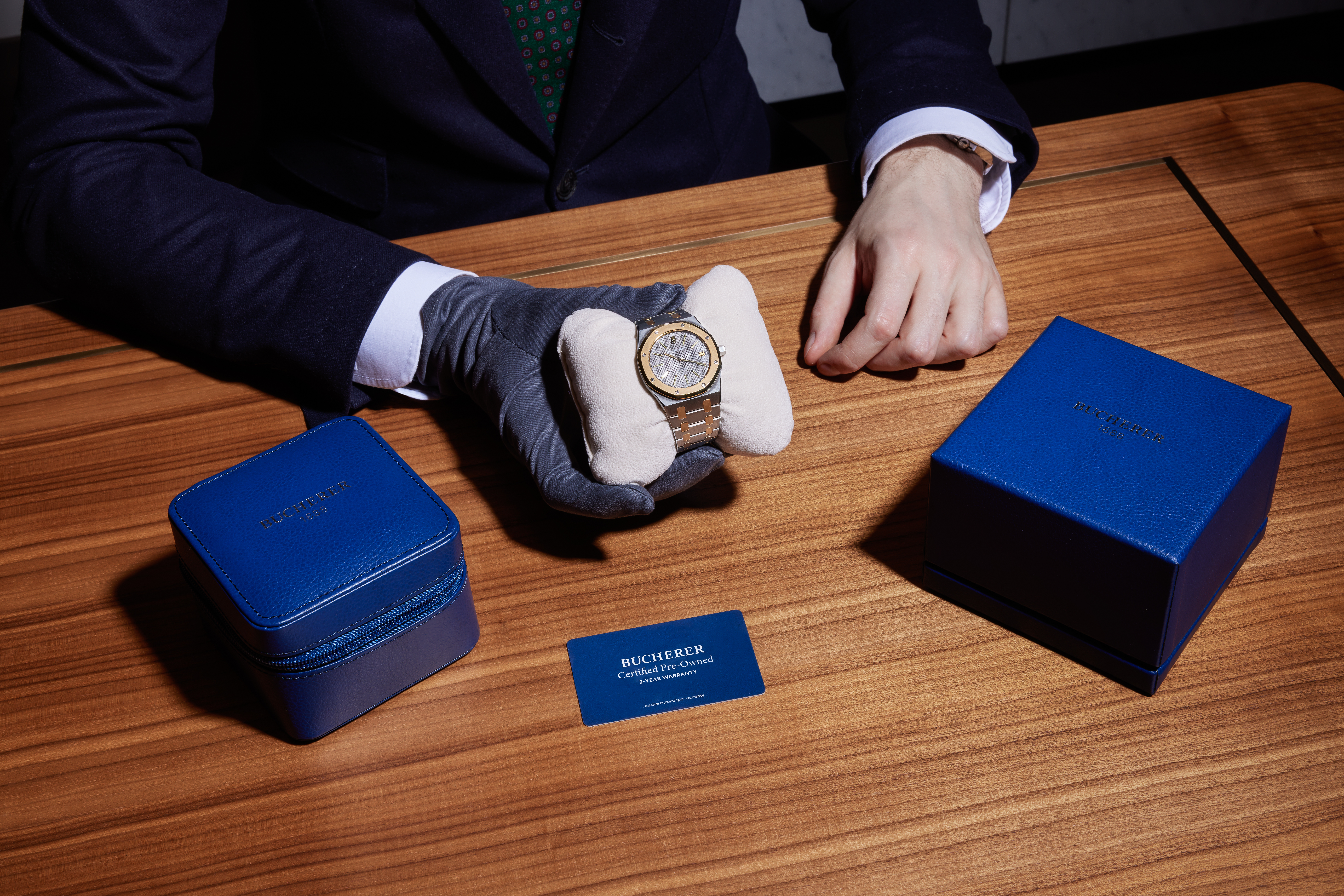 A person in a suit and gloves holds a Bucherer Certified Pre-Owned watch, with a blue box, travel case, and certificate on a wooden table.