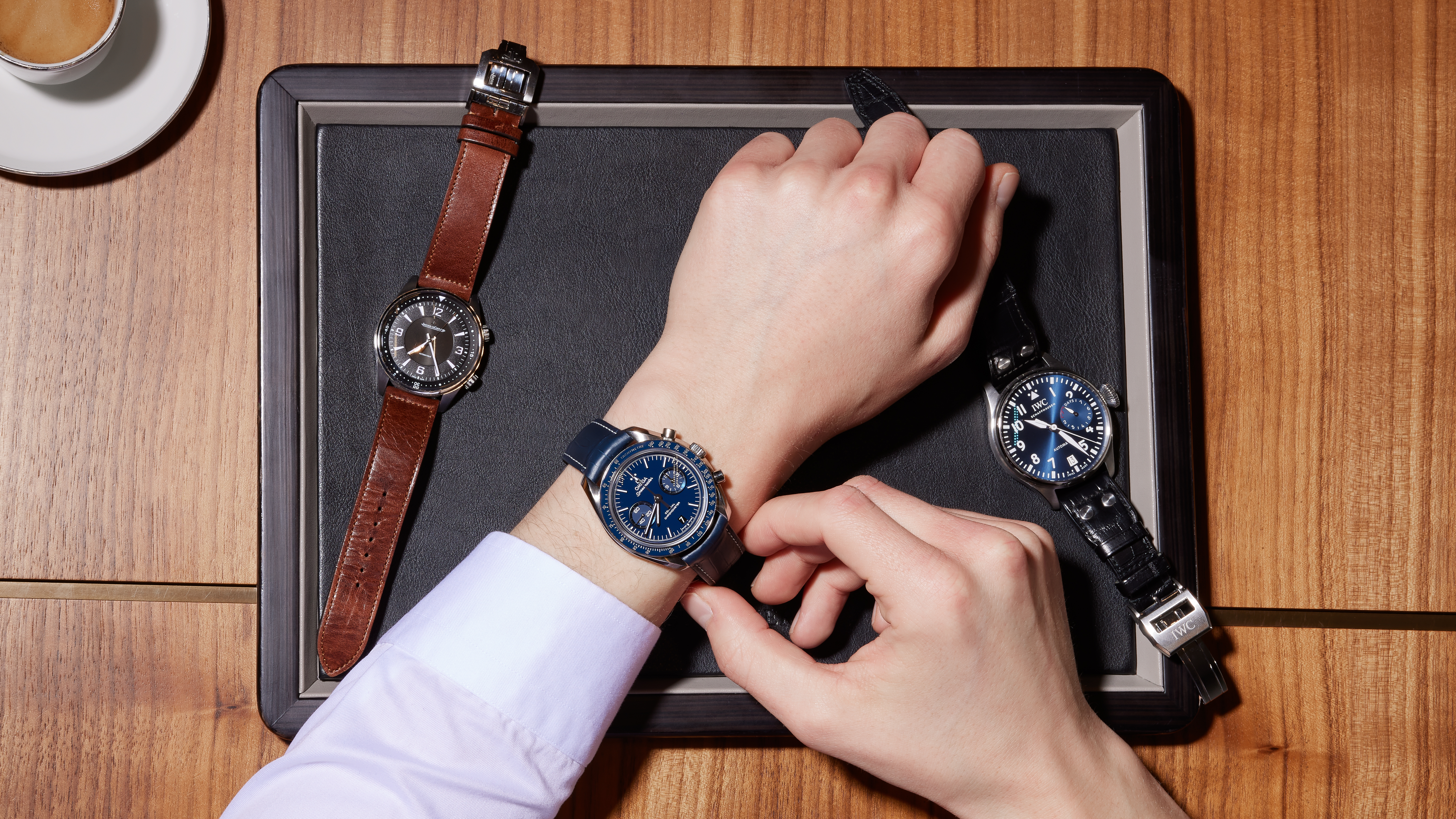 A person's hands adjusting a blue watch on their wrist, with two other watches on a tray and a cup of coffee nearby.