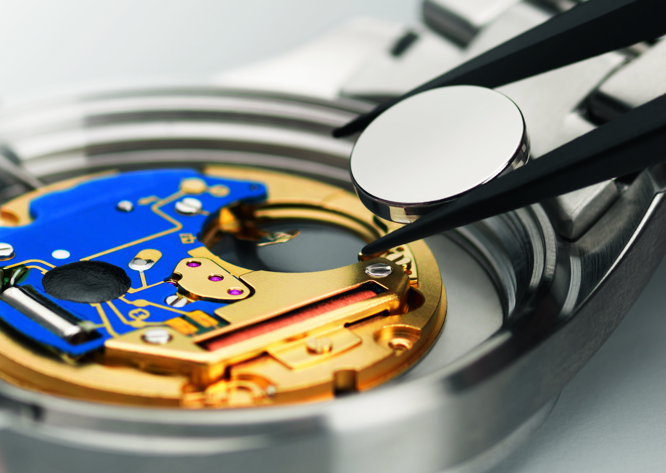 Eine Pinzette hält eine silberne Uhrenbatterie über einem geöffneten Uhrwerk mit goldenen und blauen Komponenten.