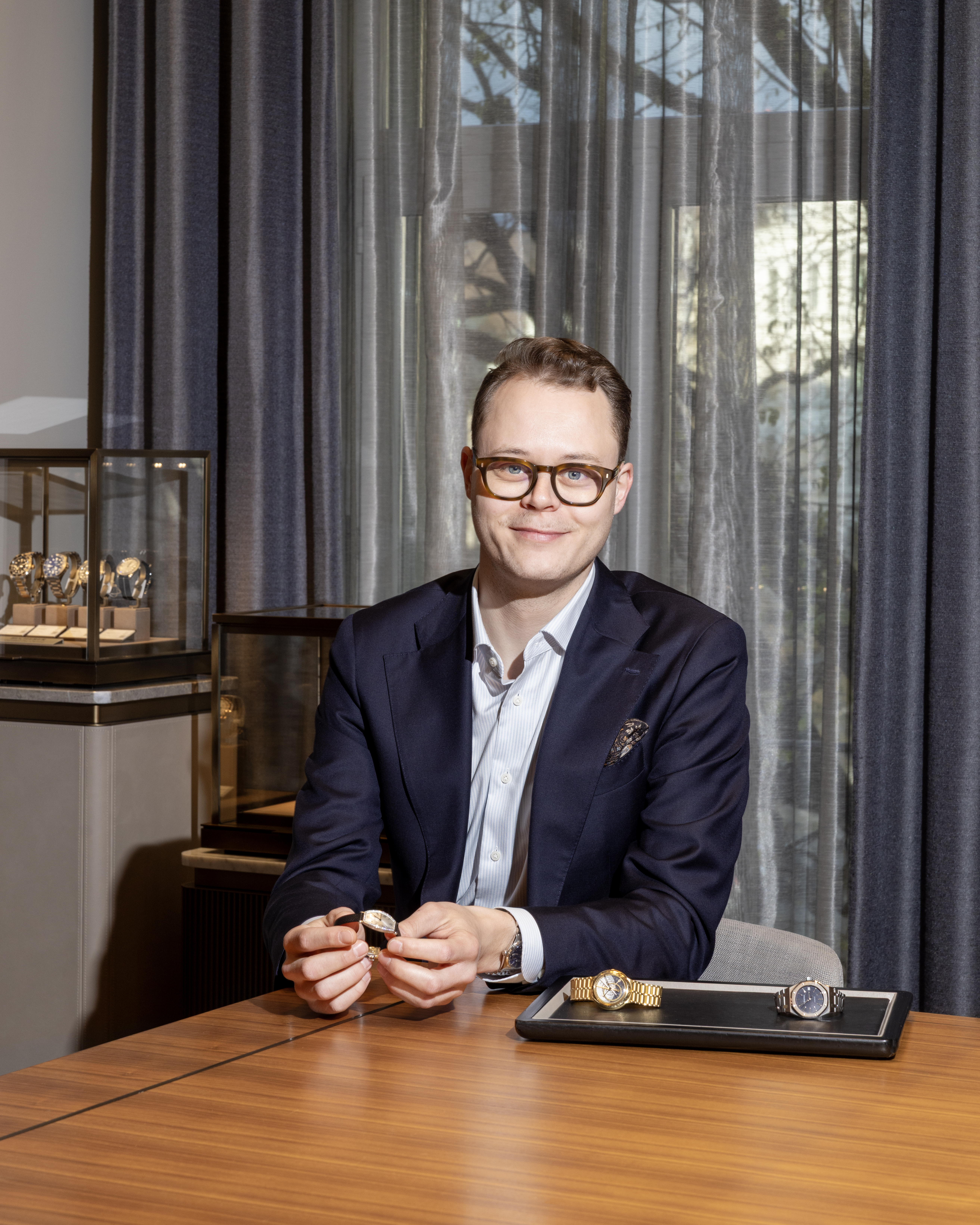 Ein Mann im Anzug und mit Brille sitzt an einem Holztisch, hält eine Uhr in der Hand, in der Nähe sind weitere Uhren ausgestellt.