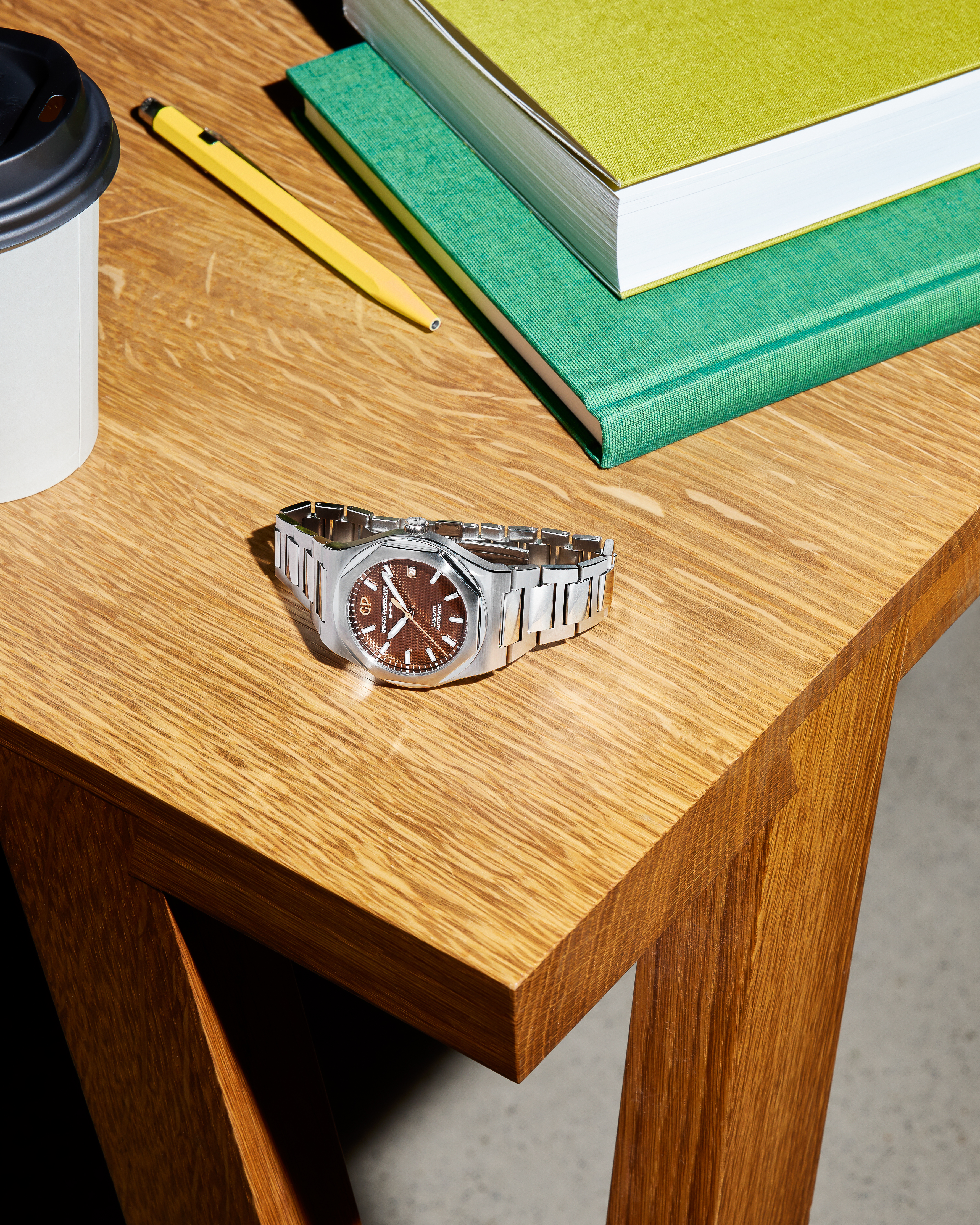 A silver watch with a brown face on a wooden desk next to a coffee cup, pencil, and two books.