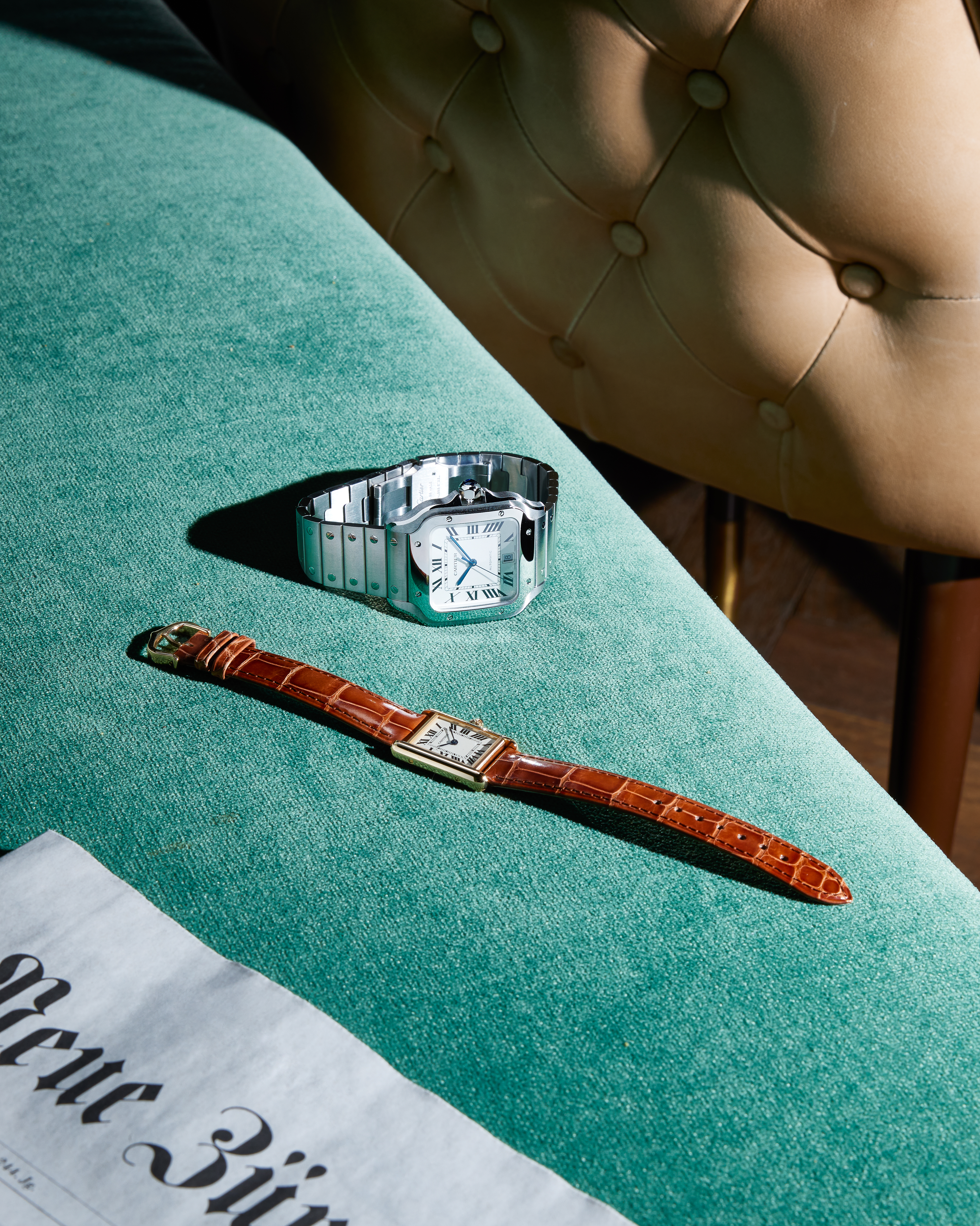 Two watches, one silver with a metal band and one gold with a brown leather strap, on a green upholstered surface.
