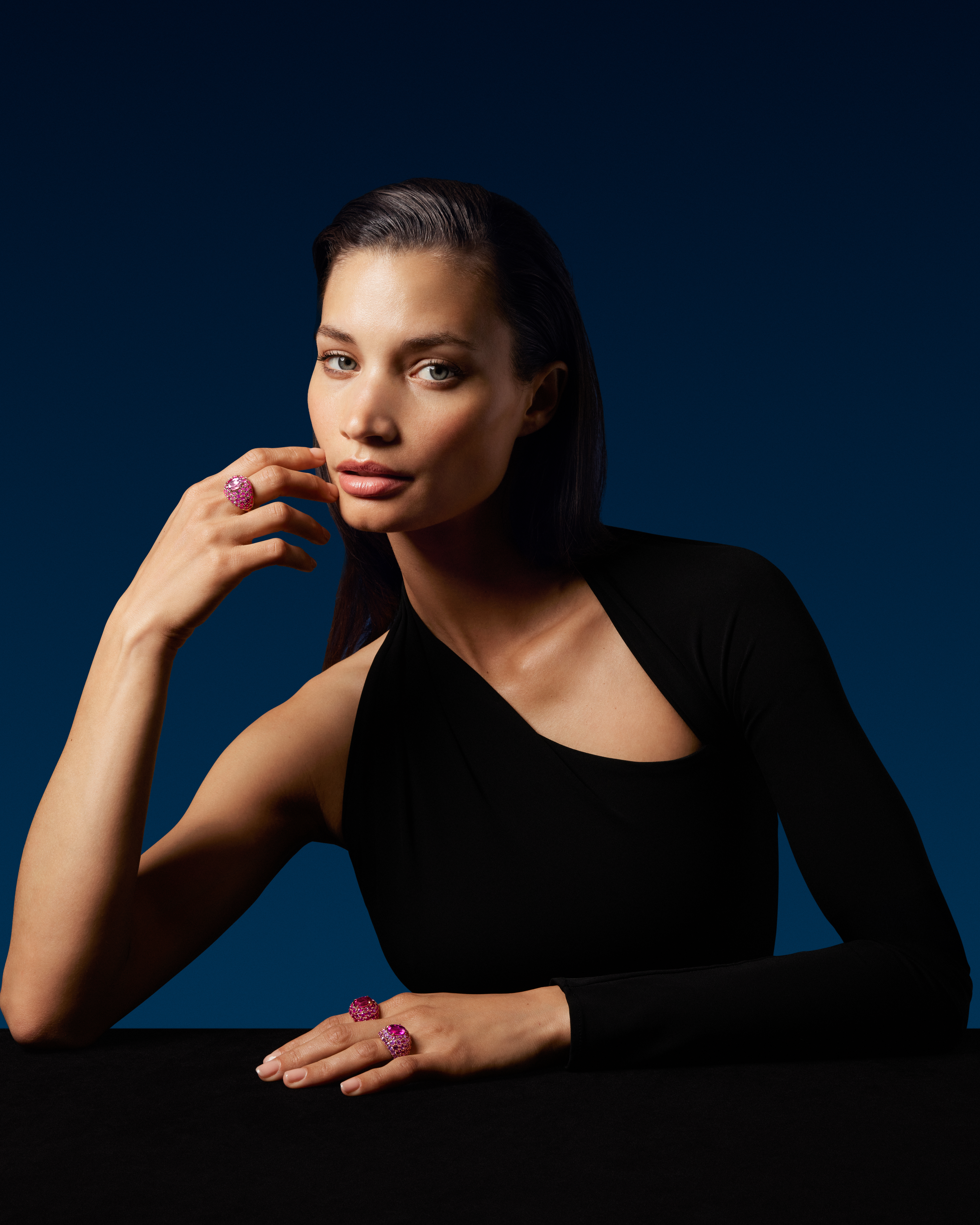 Une femme en robe noire, portant deux bagues ornées de pierres précieuses roses à la main droite, pose son bras sur une surface noire.