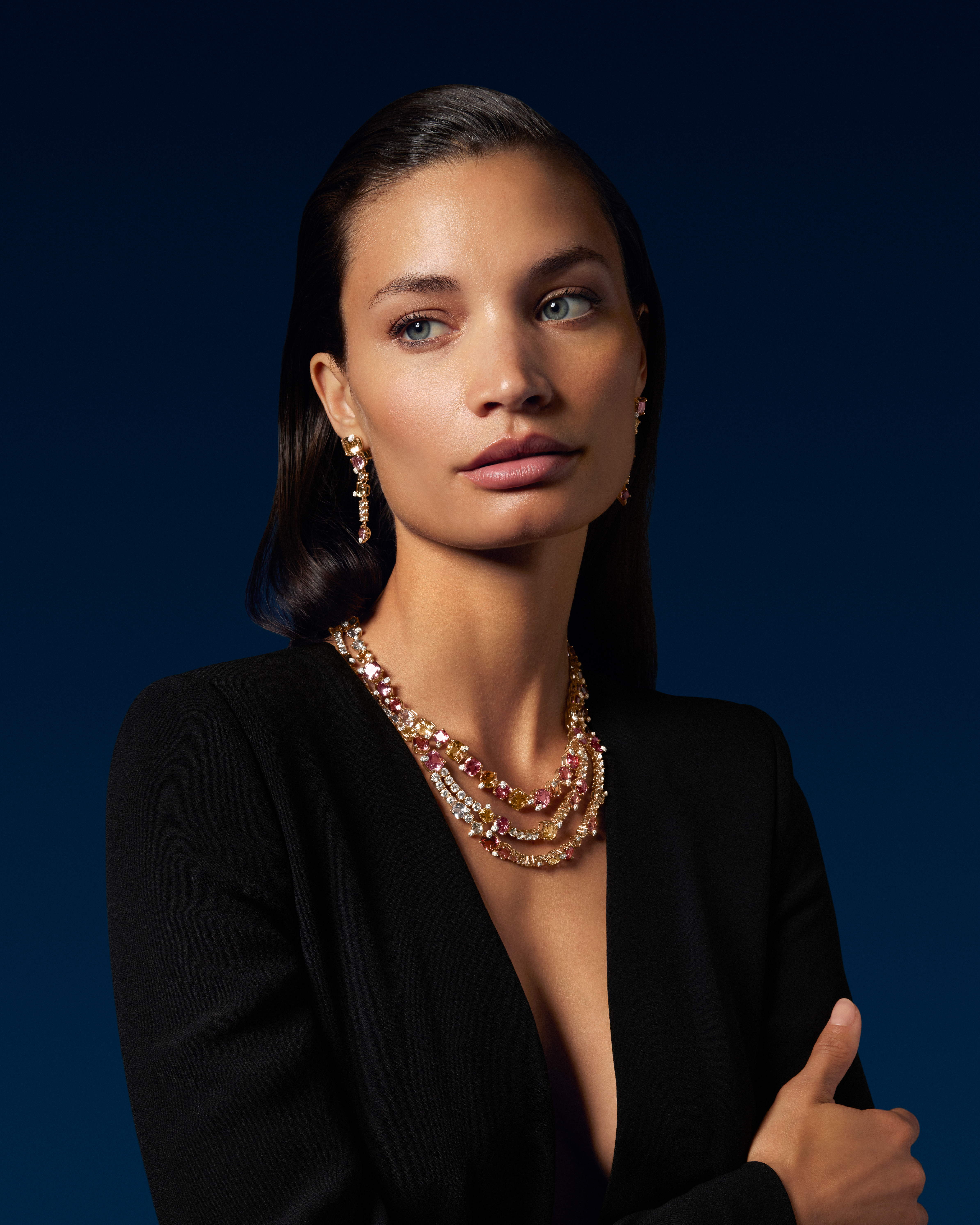 Eine Frau mit dunklem Haar und blauen Augen trägt einen schwarzen Blazer, eine mehrreihige Halskette mit rosa und gelben Edelsteinen und dazu passende Ohrringe.