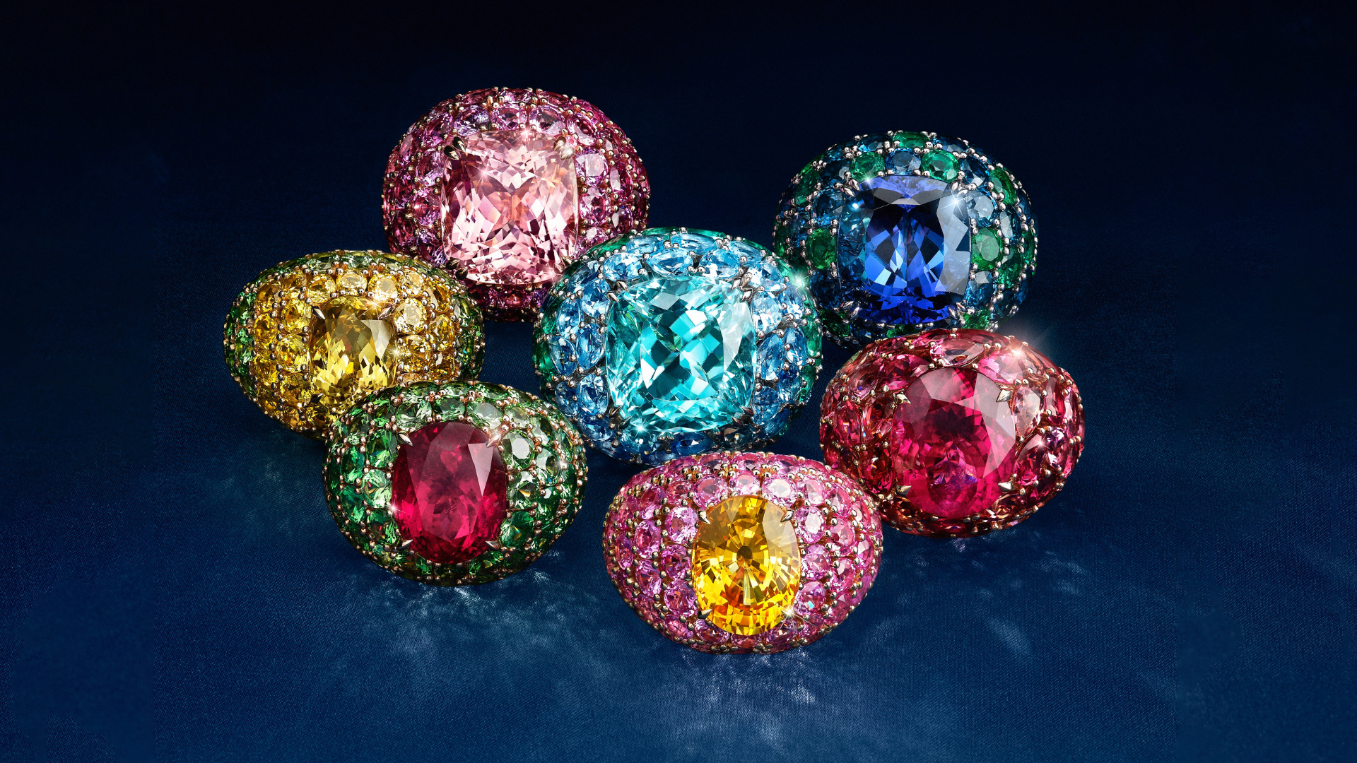 Seven colorful, ornate gemstone rings with large central stones, arranged in a cluster on a dark blue surface.