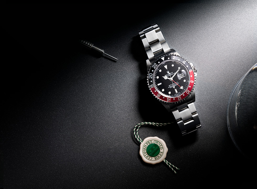 A Rolex GMT-Master II "Pepsi" watch with a steel bracelet, a spring bar tool, and a green "Certified Pre-Owned" tag on a dark surface.