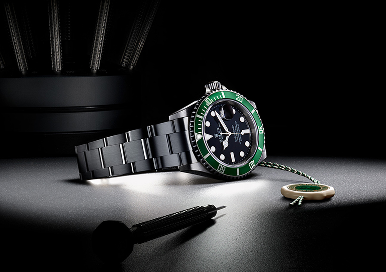 A Rolex Submariner watch with a black dial and green bezel, a dart, and a Rolex seal on a dark surface.