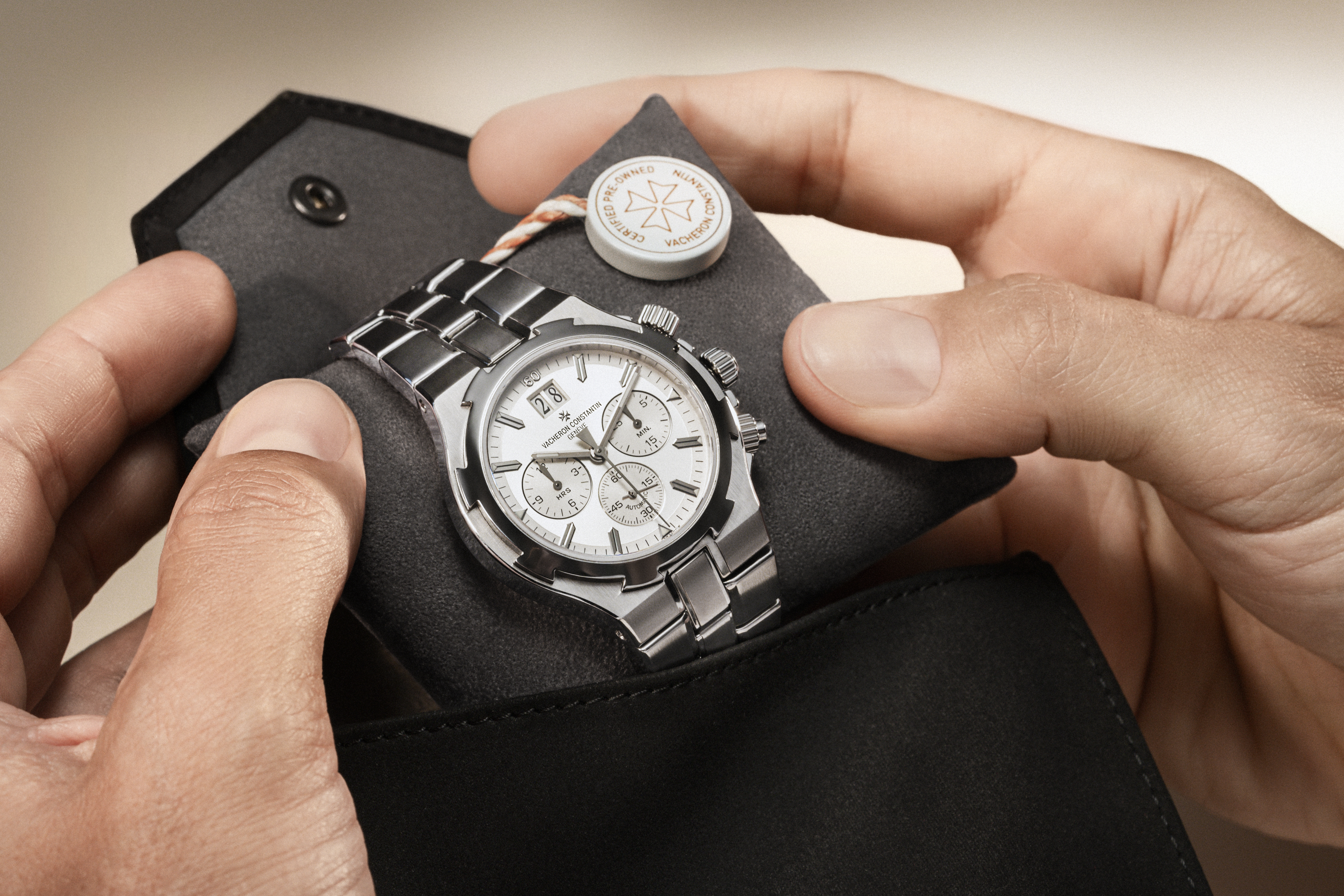Hands hold a silver chronograph watch with a white face and metal band, resting on a dark gray cushion inside a black case.