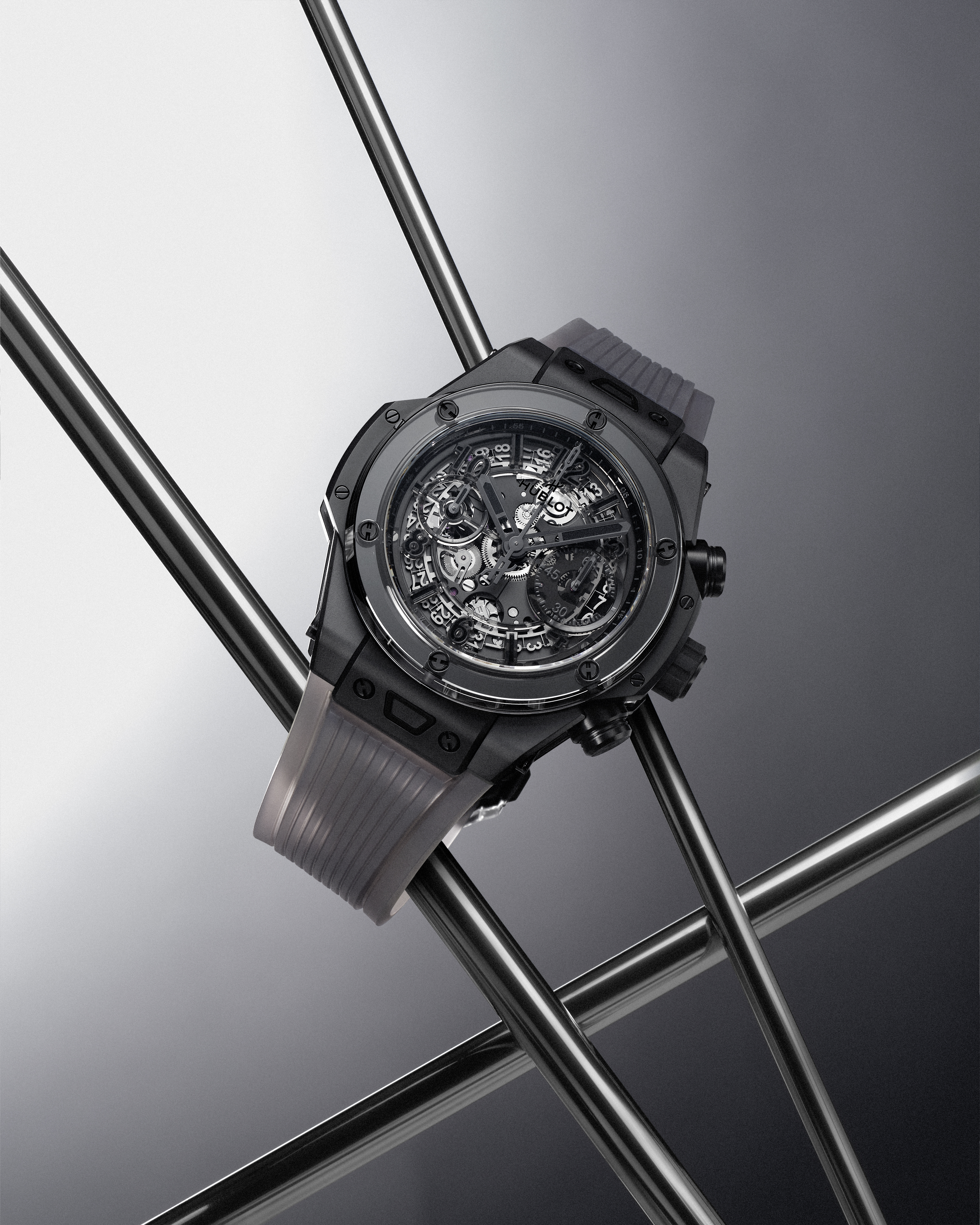 A black skeleton watch with a translucent gray strap rests on intersecting metal rods against a gradient gray background.