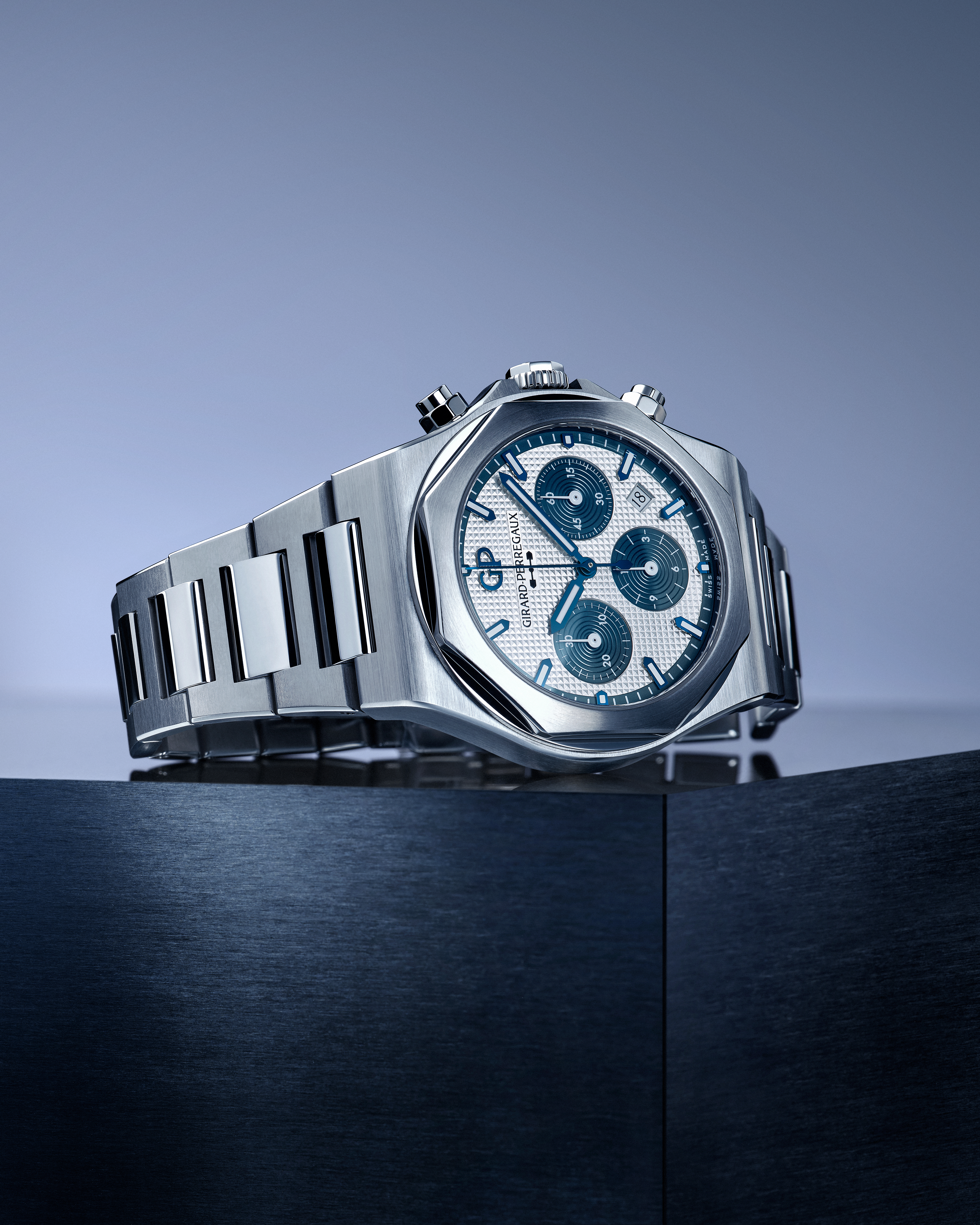 A silver chronograph watch with a white dial and blue accents, resting on a dark blue surface.