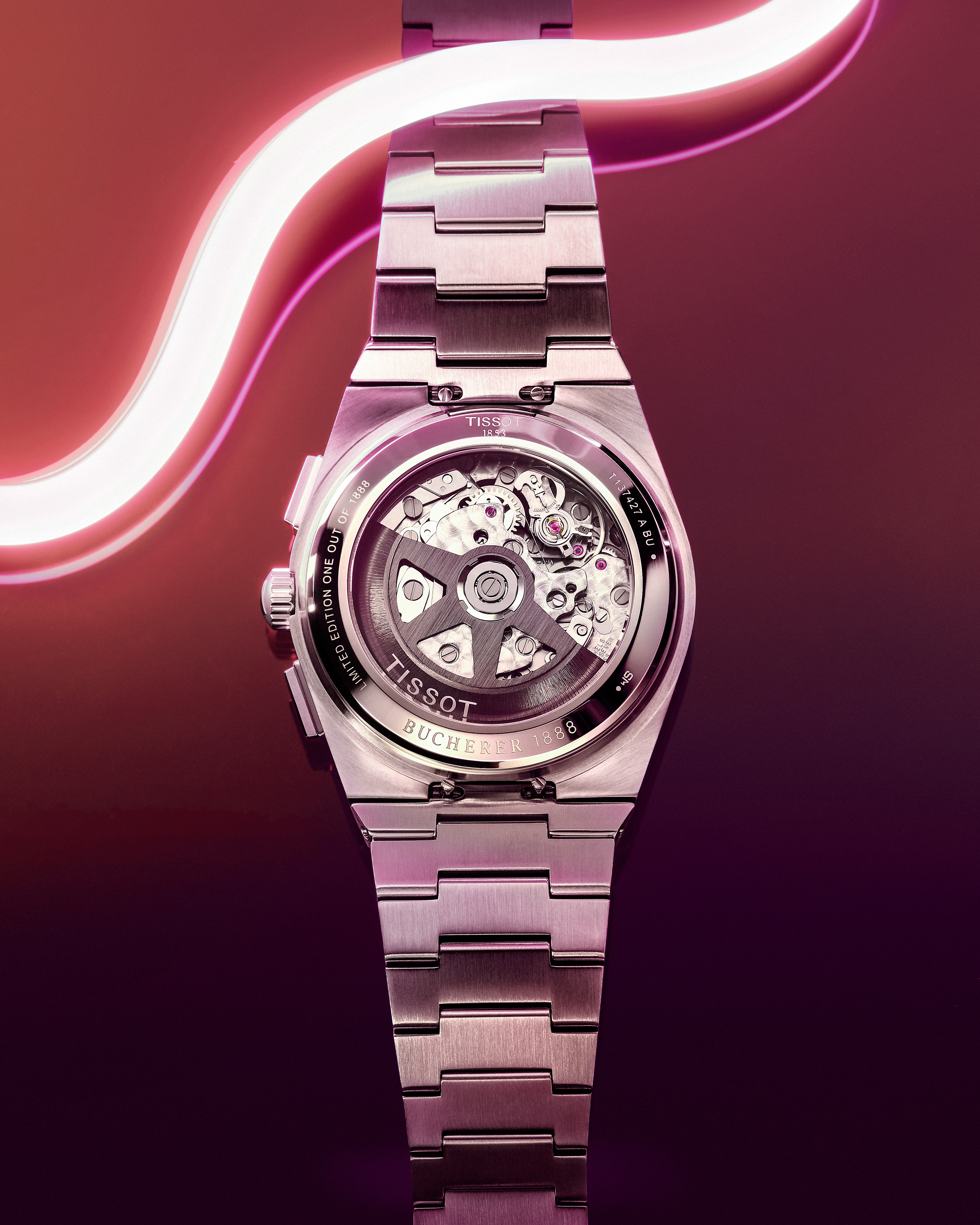 The back of a silver Tissot watch with a visible mechanical movement, illuminated by a pink neon light.