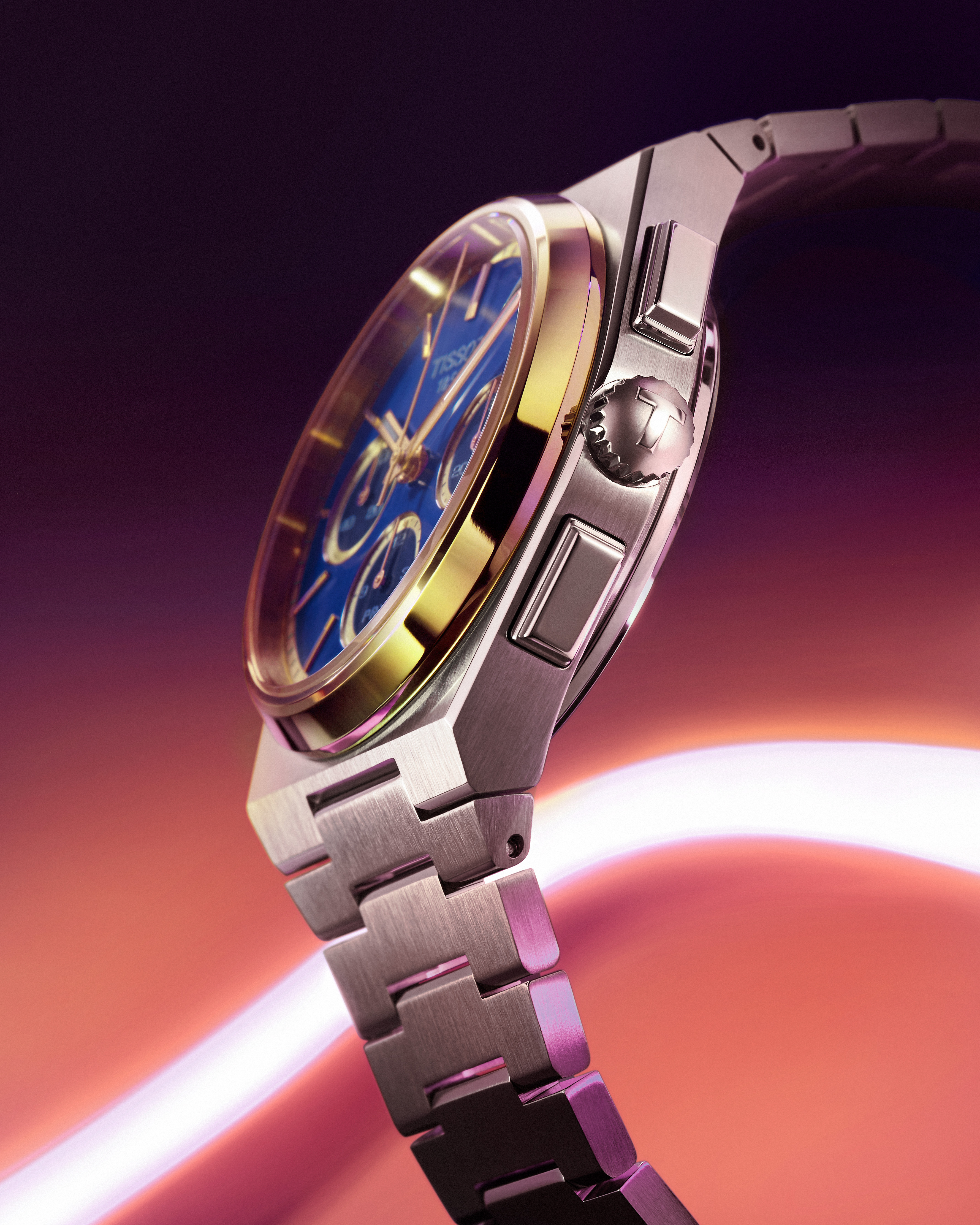 A silver and gold Tissot watch with a blue face and three subdials, angled against a blurred purple and orange background.
