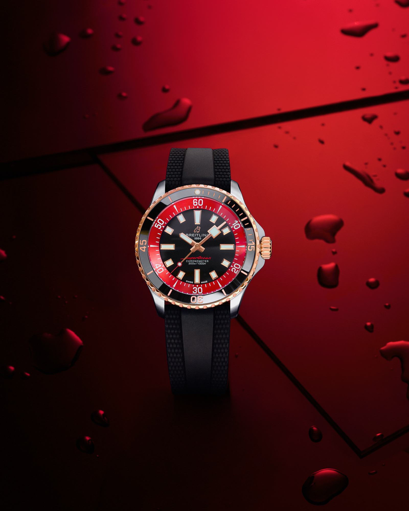 Breitling Superocean watch with a black dial, red and gold bezel, and black rubber strap on a red surface with water droplets.