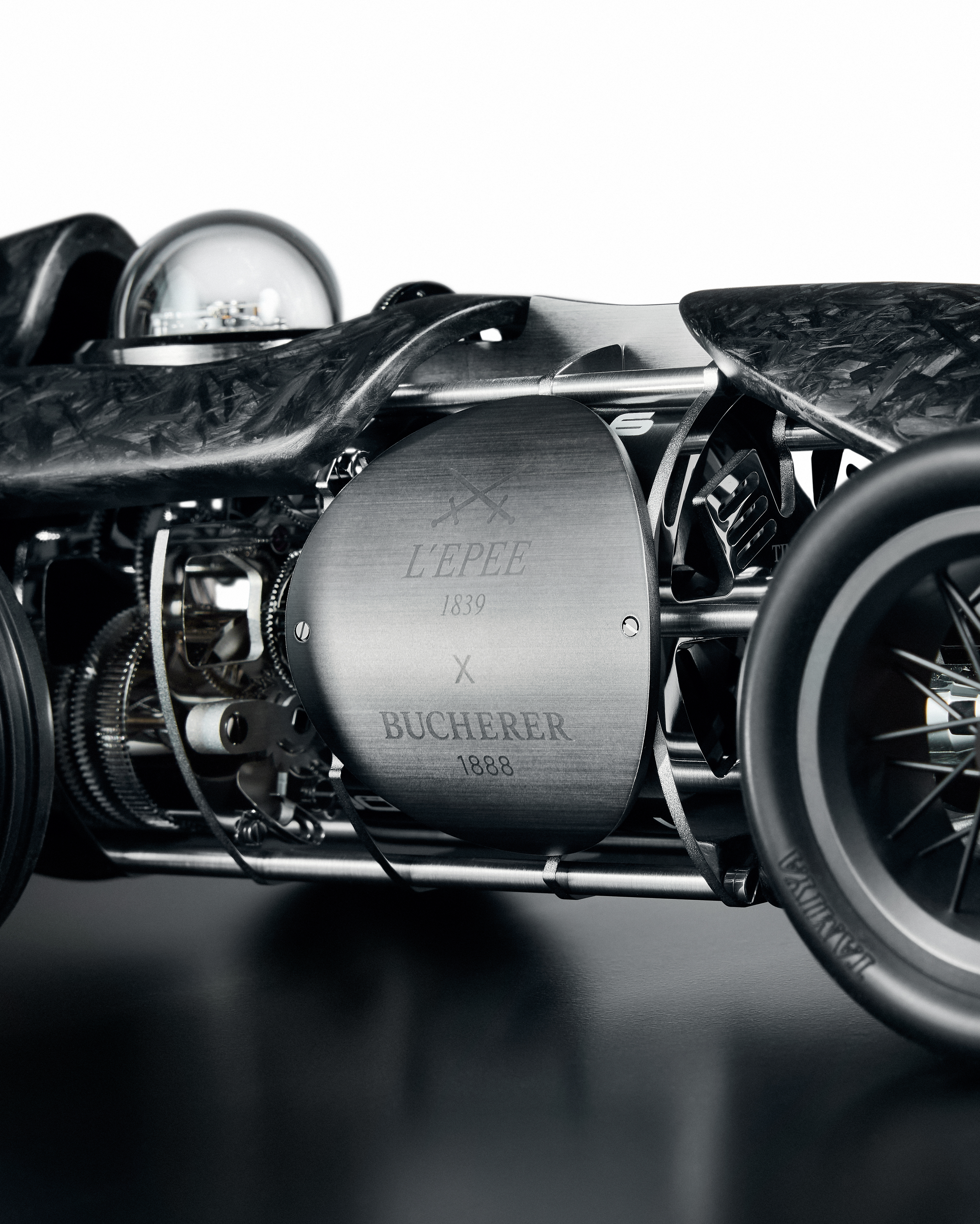 Gros plan sur une voiture de course miniature, complexe, gris foncé, avec des engrenages visibles et un panneau latéral gravé de « L'EPEE 1839 X BUCHERER 1888 ».