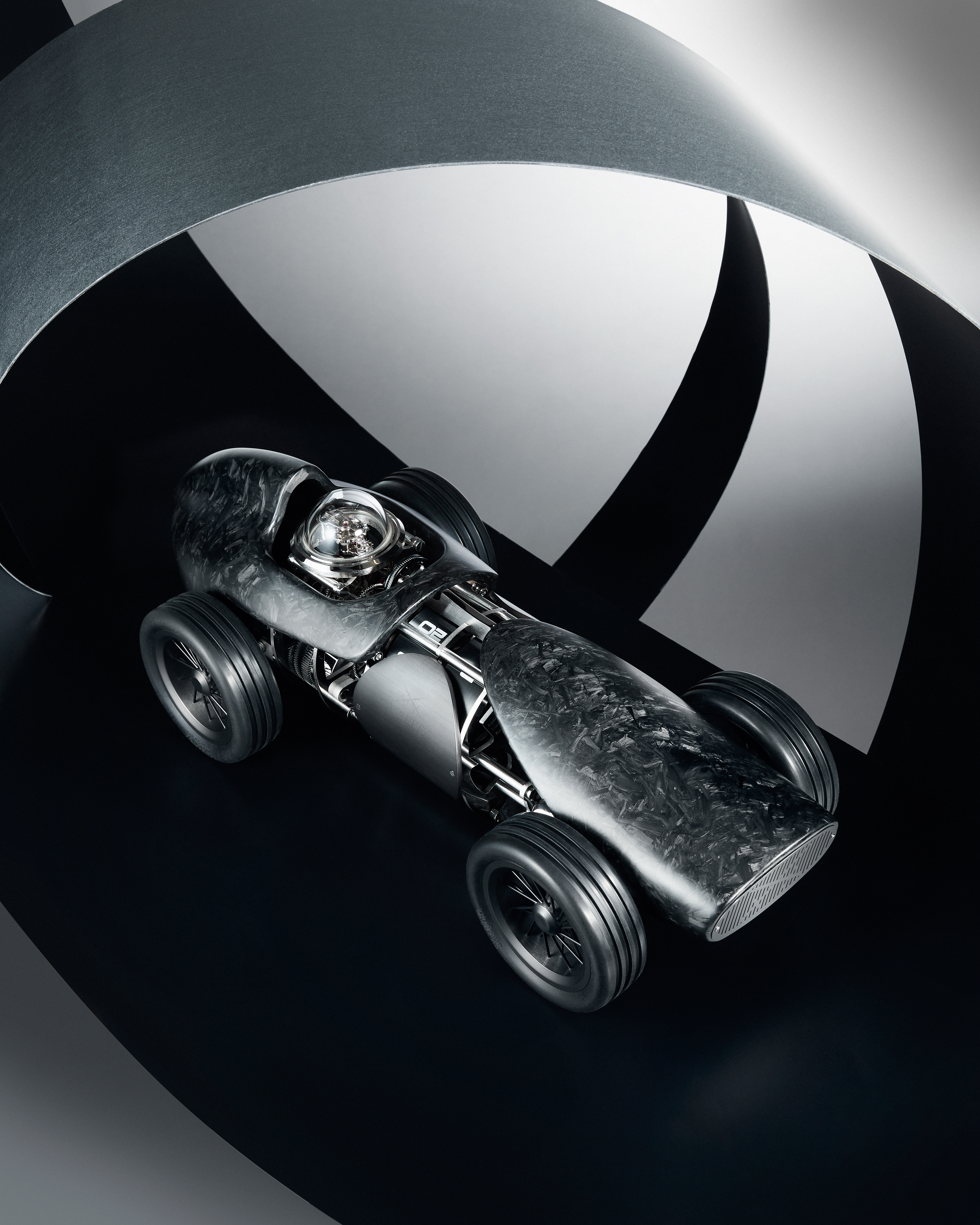 A sleek, dark gray, carbon fiber race car model with exposed engine components, on a black and white curved track.