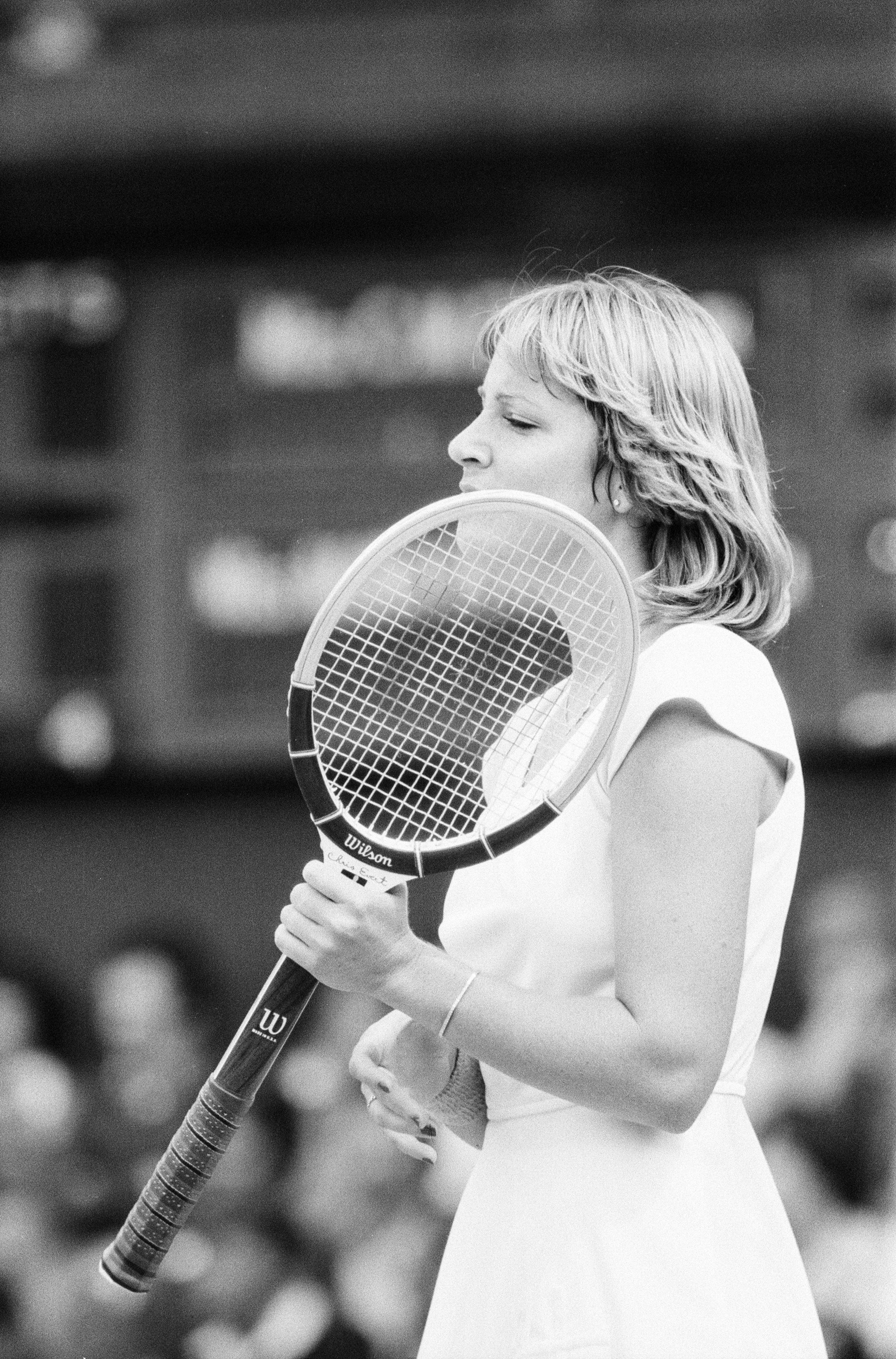 A female tennis player in a white dress holds a Wilson racket to her face, looking left.