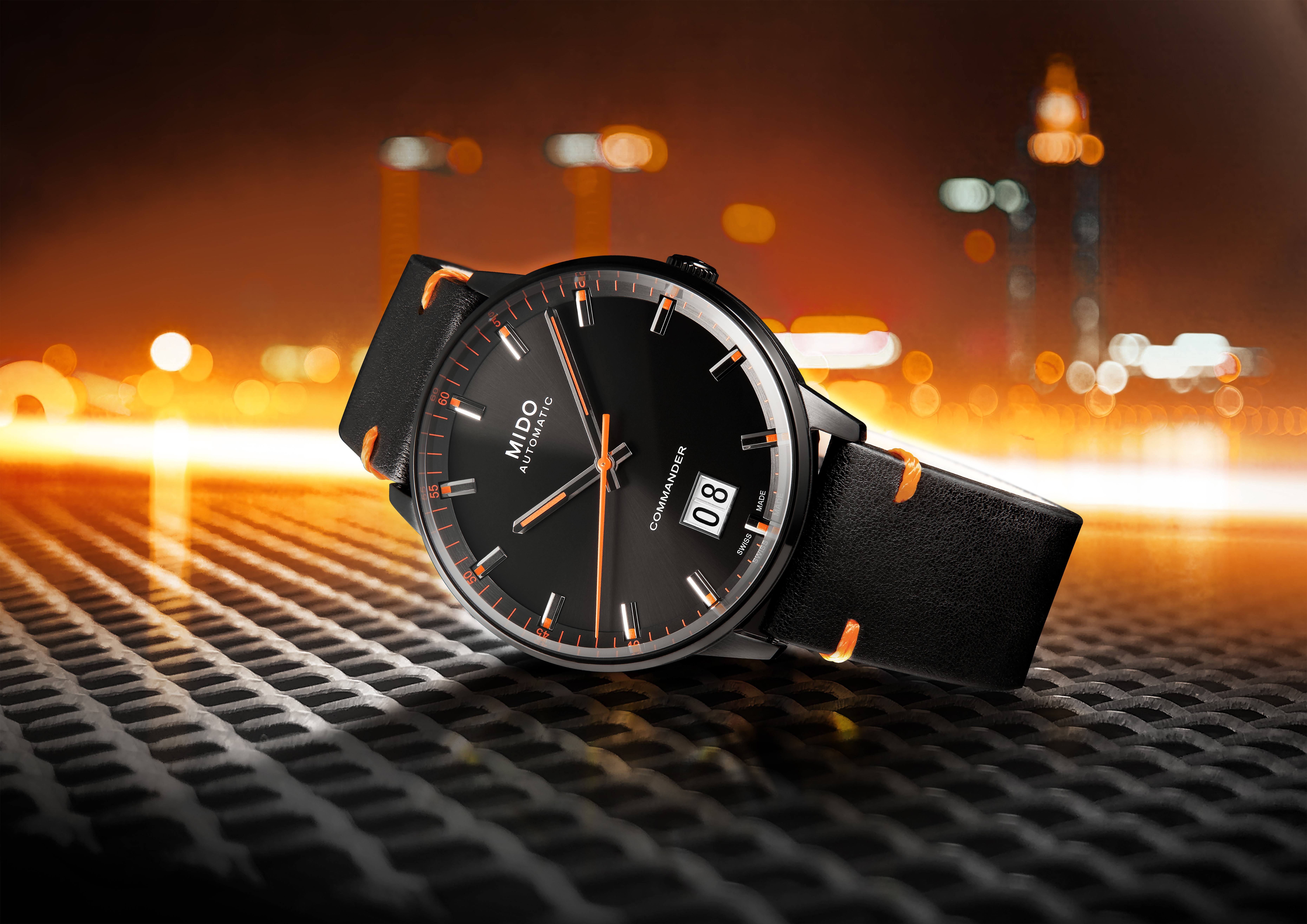 Eine schwarze Mido Commander Automatikuhr mit orangefarbenen Akzenten und schwarzem Lederarmband, die auf einem Metallgitter ruht, im Hintergrund sind verschwommene Stadtlichter zu sehen.