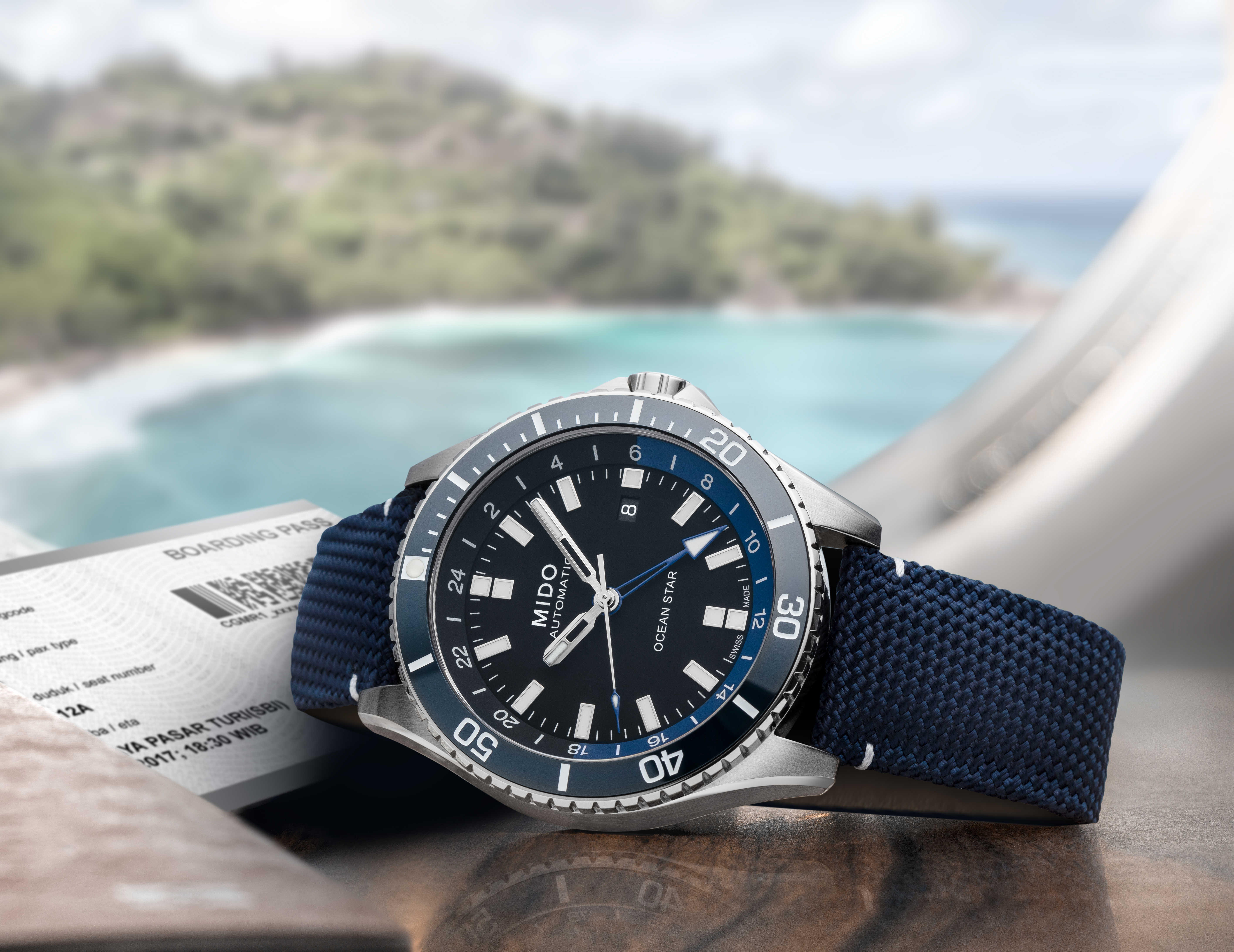 Eine Mido Ocean Star mit schwarzem Zifferblatt und blauem Stoffarmband liegt auf einer Stein-Oberfläche, neben ihr ist ein Boarding Pass zu erkennen, im Hintergrund das Meer und die Küste.