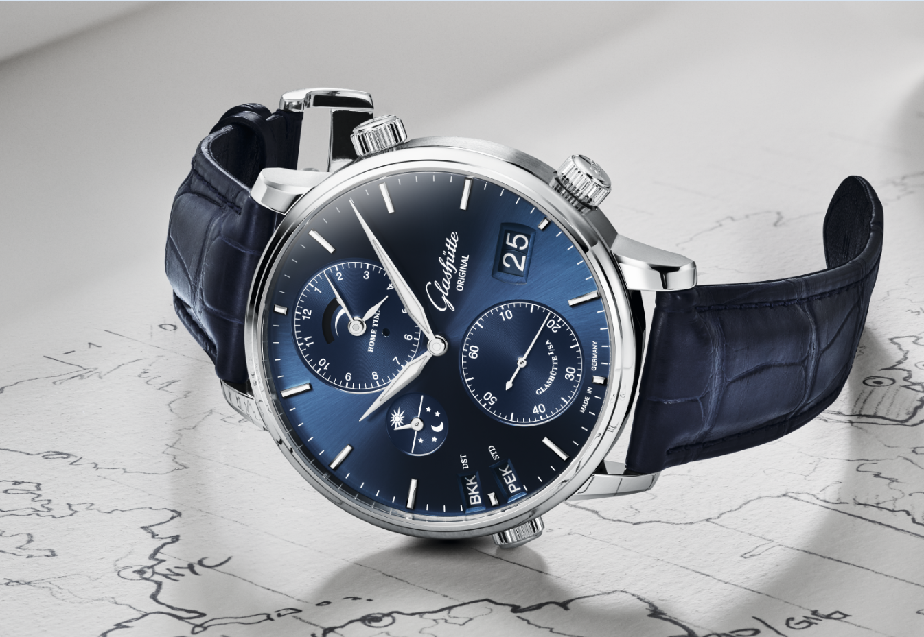 A silver Glashütte Original Senator Cosmopolite watch with a blue dial and blue alligator leather strap, resting on a map.