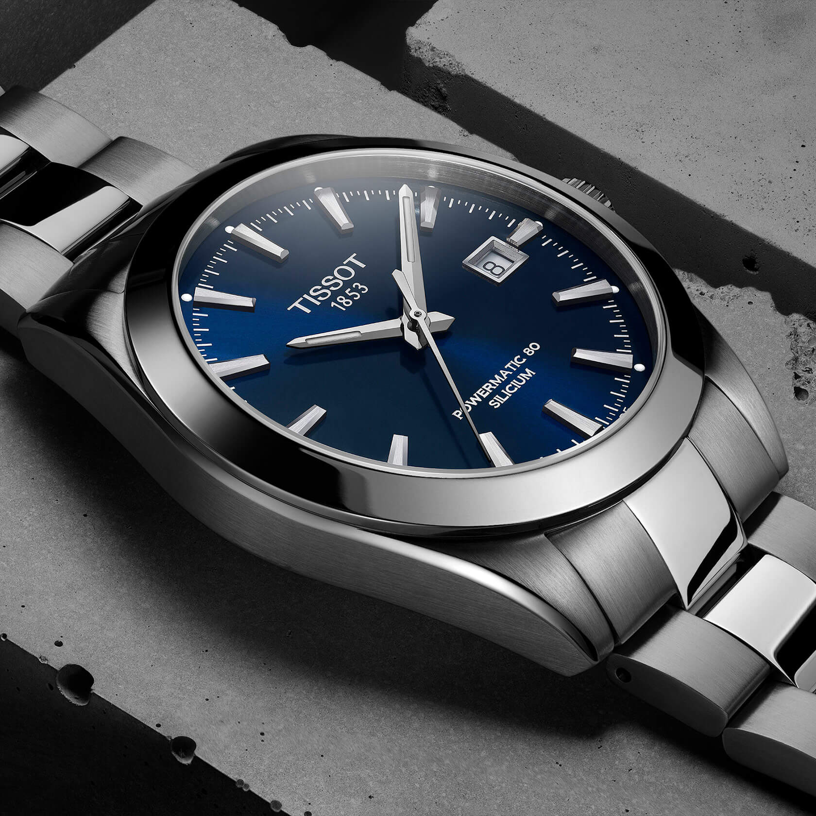 Gros plan sur une montre Tissot argentée à cadran bleu et guichet de date, posée sur une surface en béton.