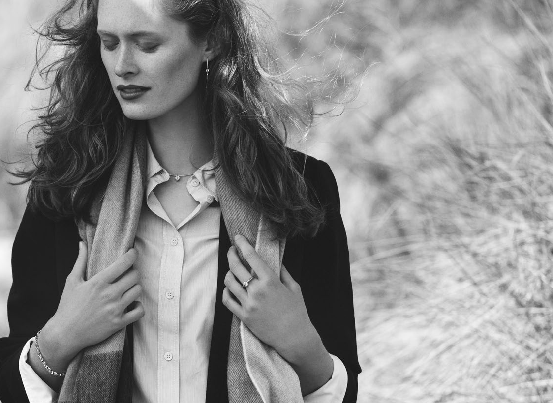 A woman with closed eyes and wind-blown hair wears a scarf, button-down shirt, jacket, and jewelry.