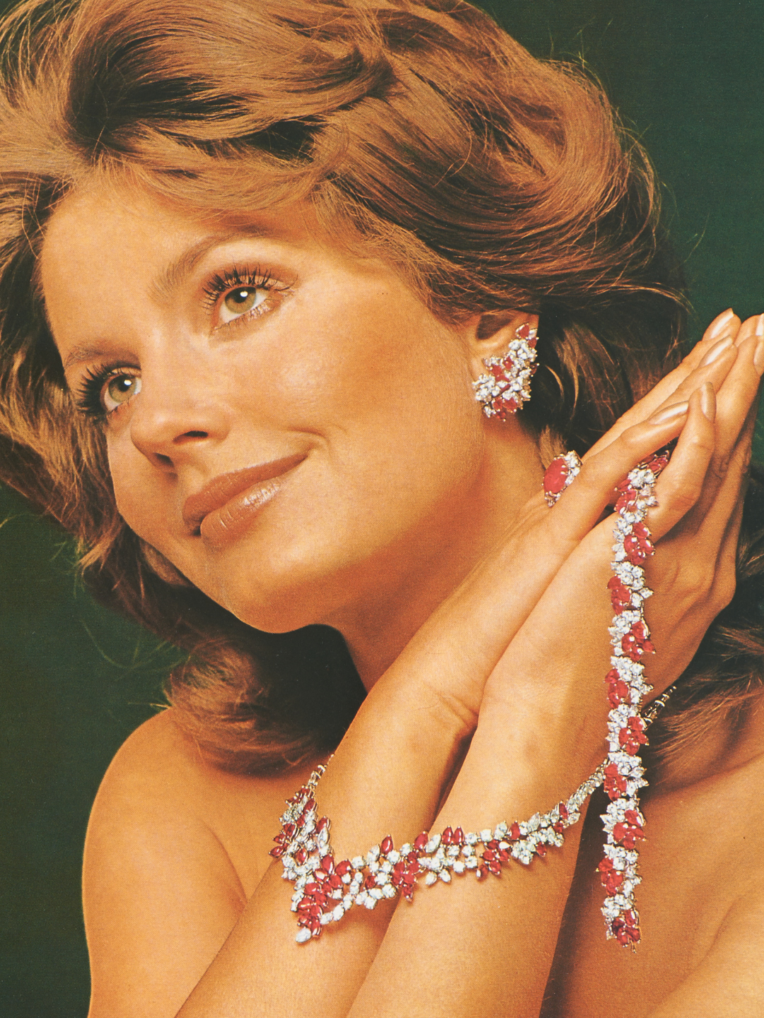 A woman with auburn hair and green eyes wears a ruby and diamond necklace, bracelet, and earrings.