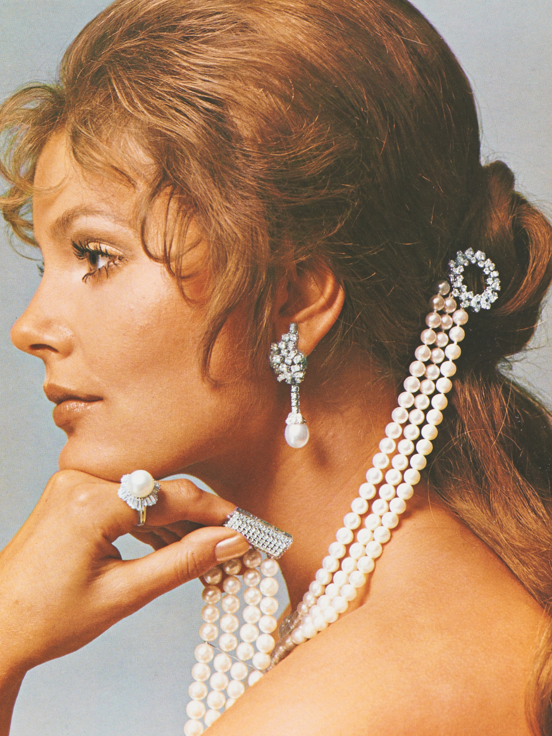 A woman in profile wears pearl and diamond earrings, a pearl ring, a diamond bracelet, and pearl strands in her hair.