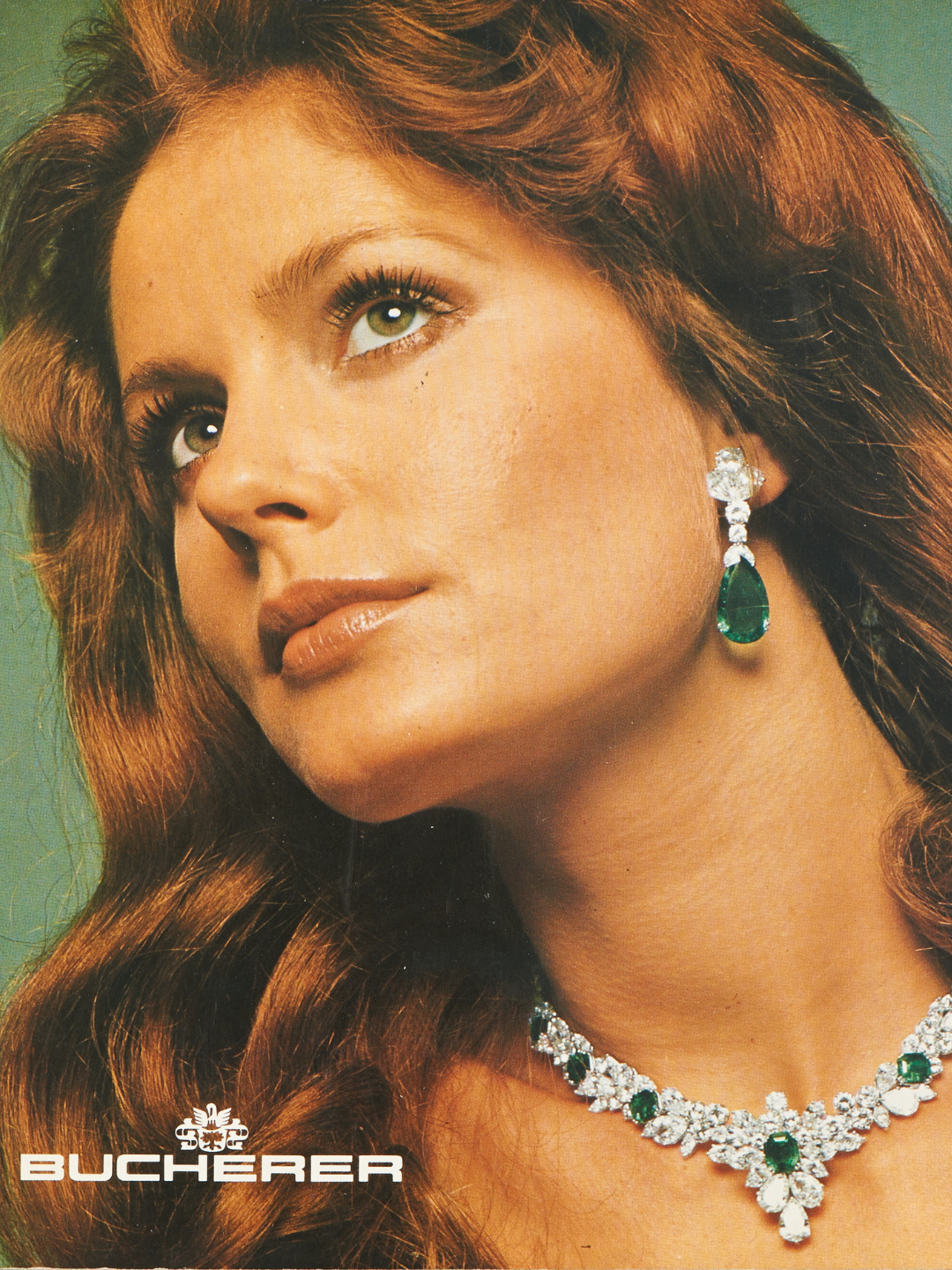 A woman with long auburn hair wears a diamond and emerald necklace and matching drop earring.